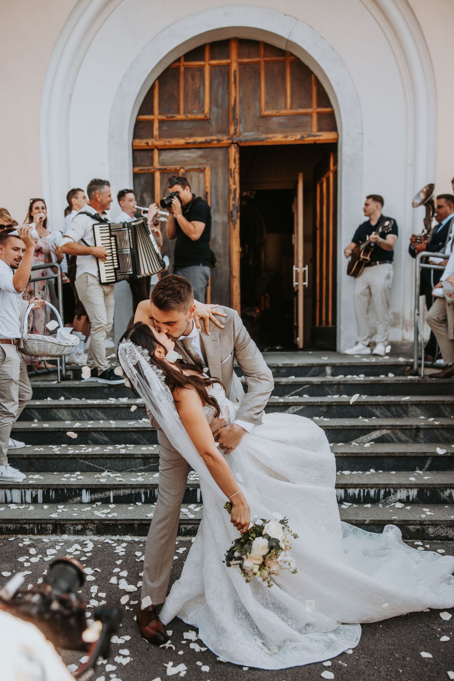 Groom dips bride for romantic kiss on church steps while musicians play and confetti falls.