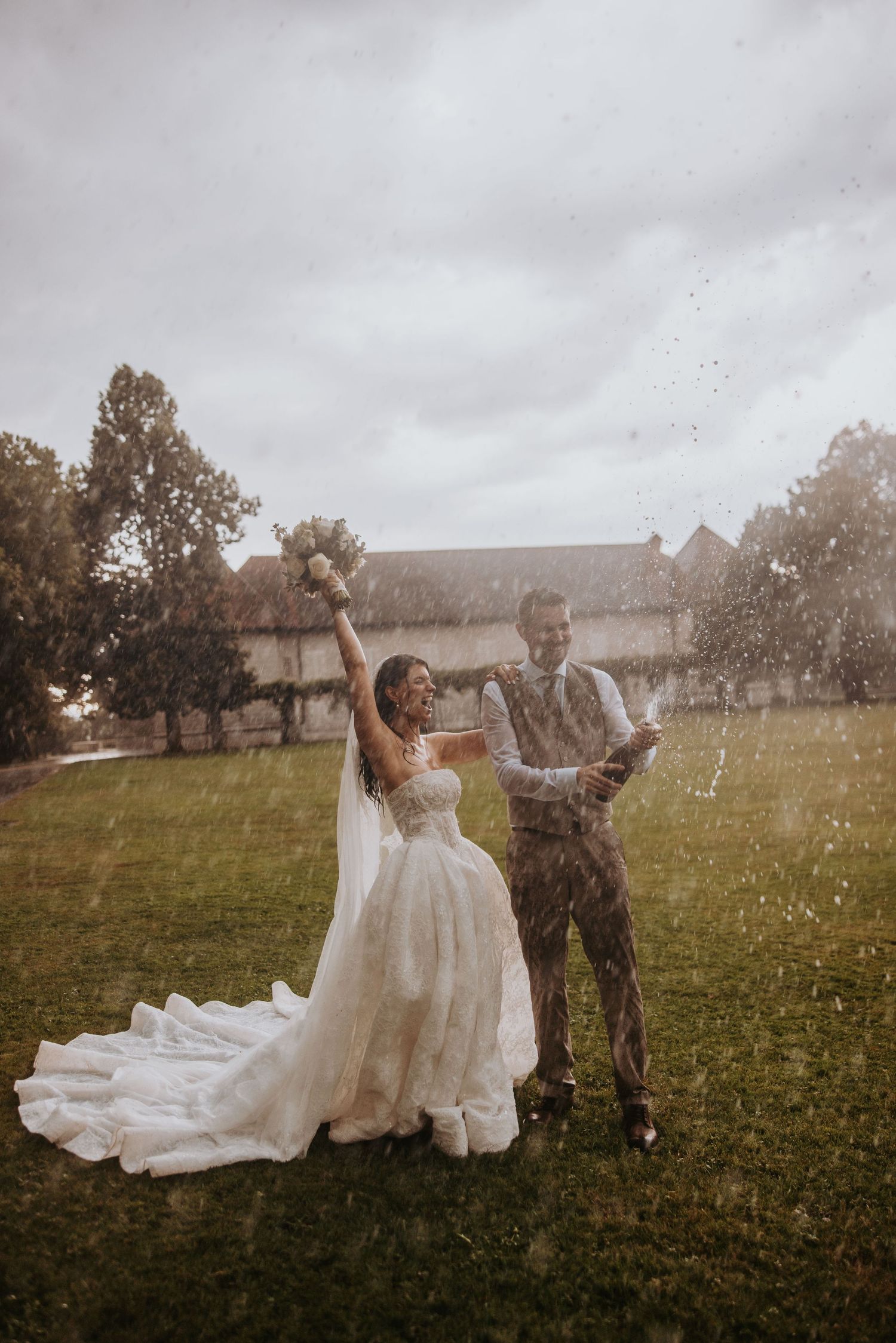 Joyful newlyweds popping champagne in the rain, bride raising bouquet, groom in grey suit on green lawn.