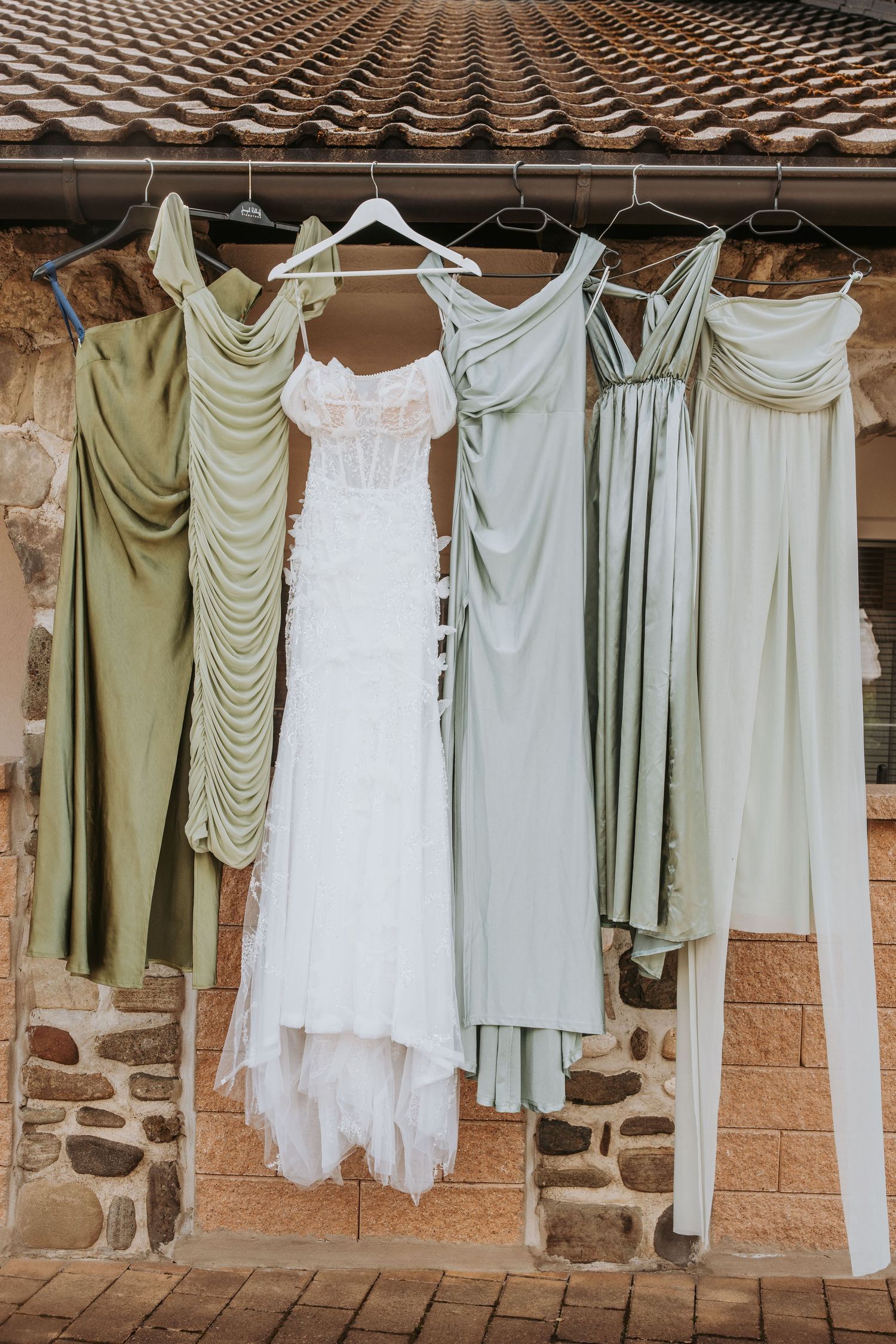 Wedding gown and sage green bridesmaid dresses hanging outdoors against a rustic stone wall.