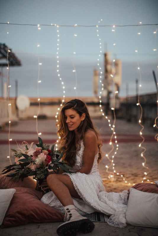 Bride in lace dress sitting with bouquet surrounded by fairy lights.