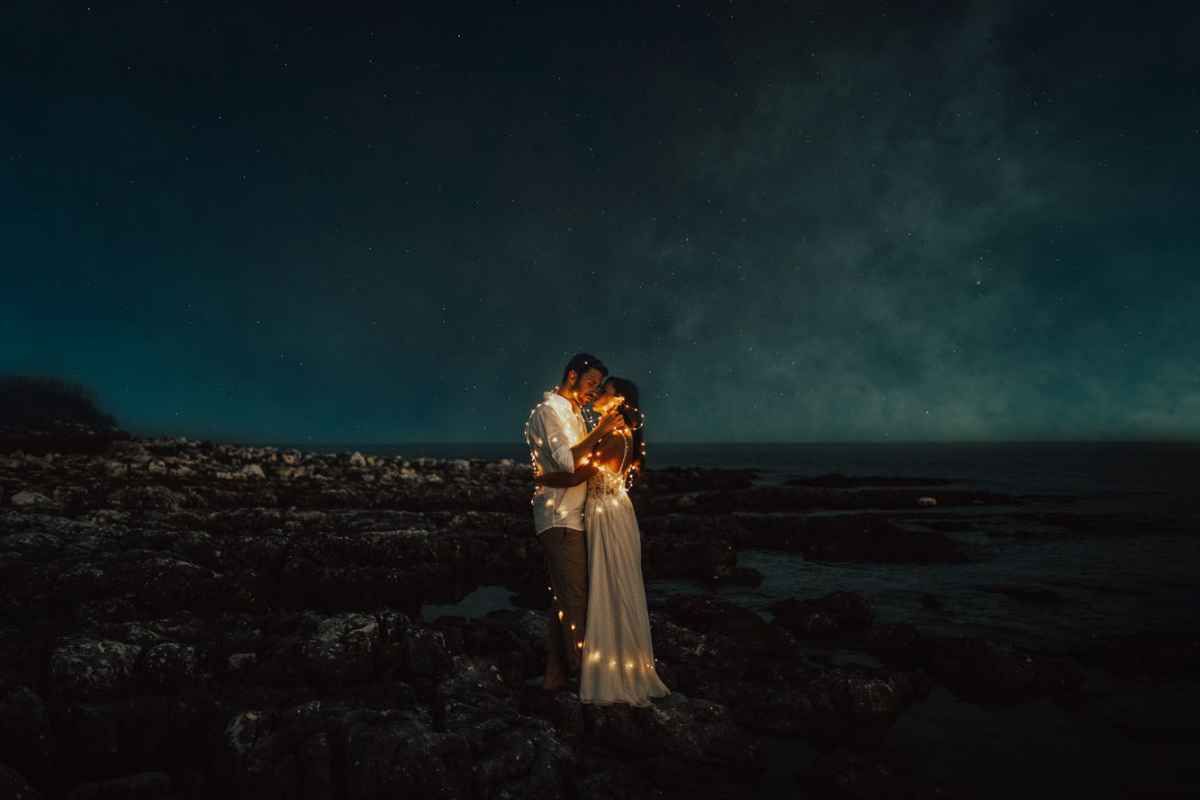 Couple embracing under a starry night sky on a rocky beach, illuminated by warm string lights.