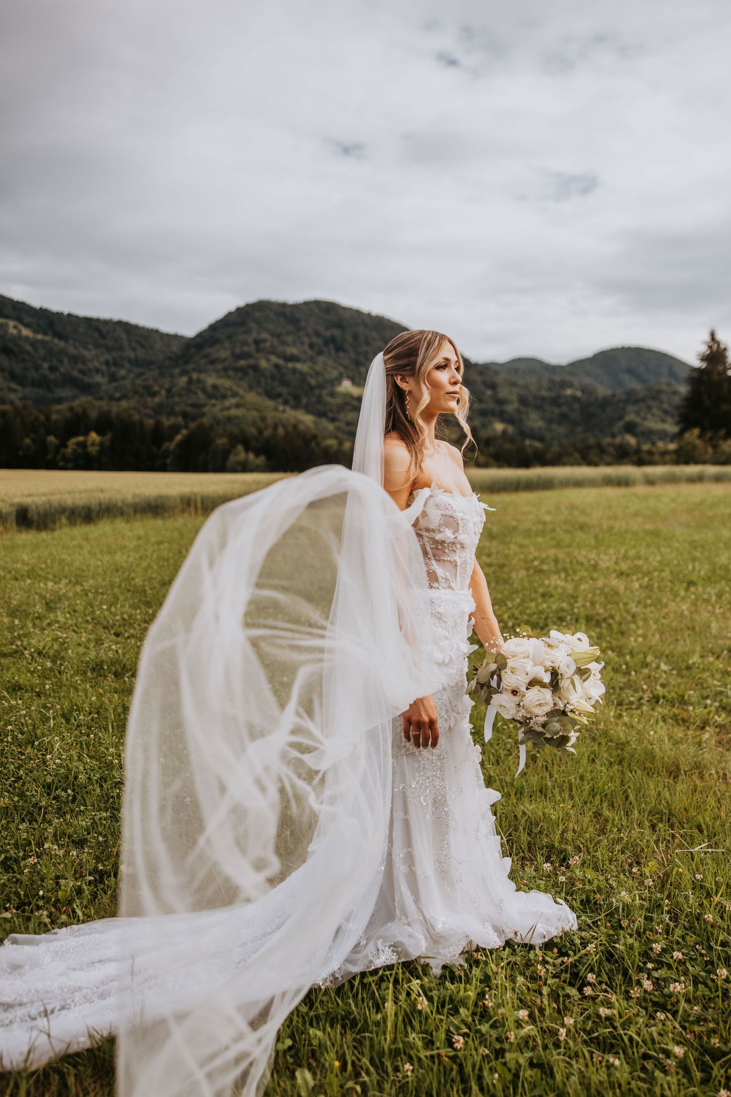 Bride in lace gown with flowing veil standing in green meadow with mountain backdrop.
