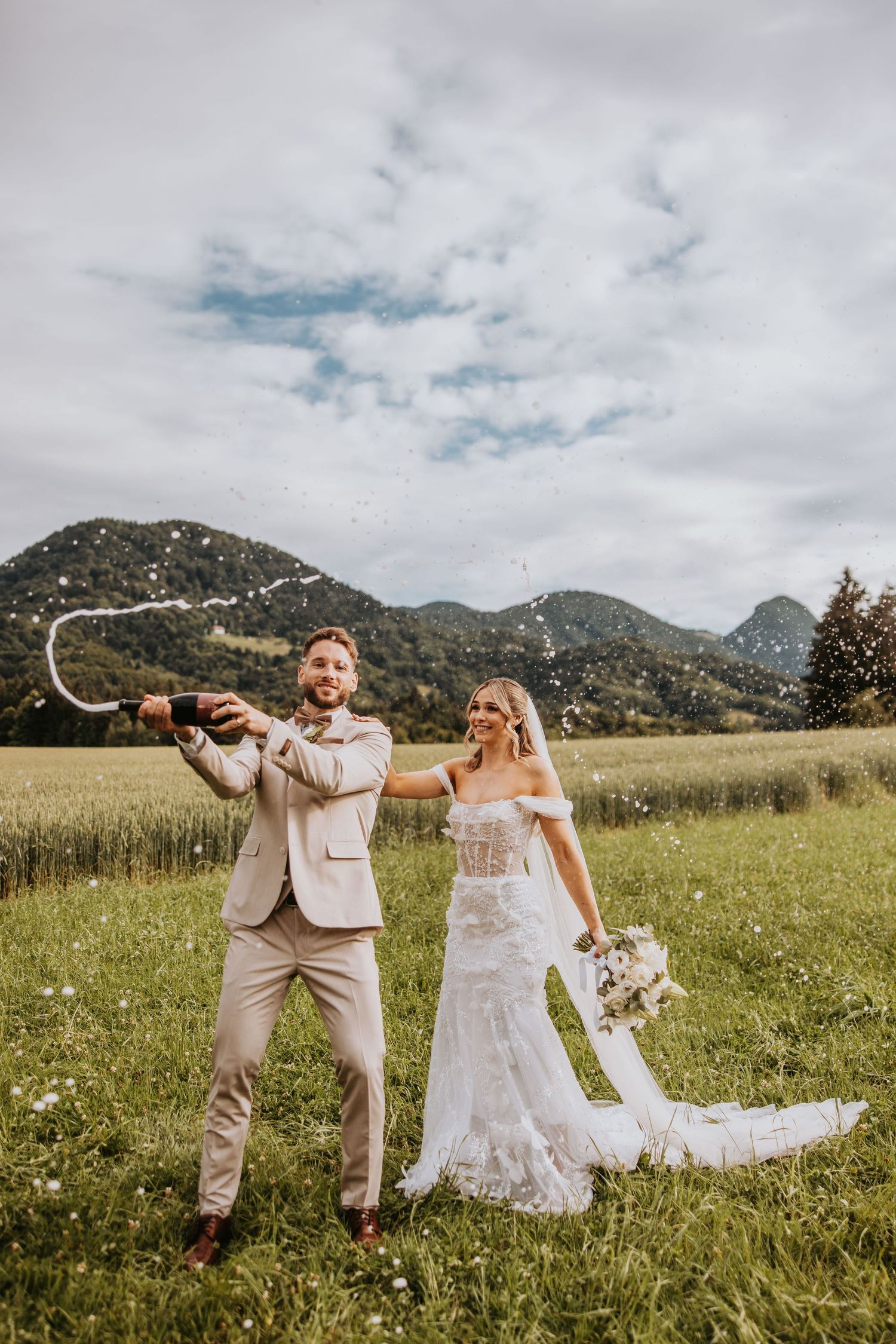 Bride and groom celebrate with champagne spray in a scenic mountain meadow on their wedding day.