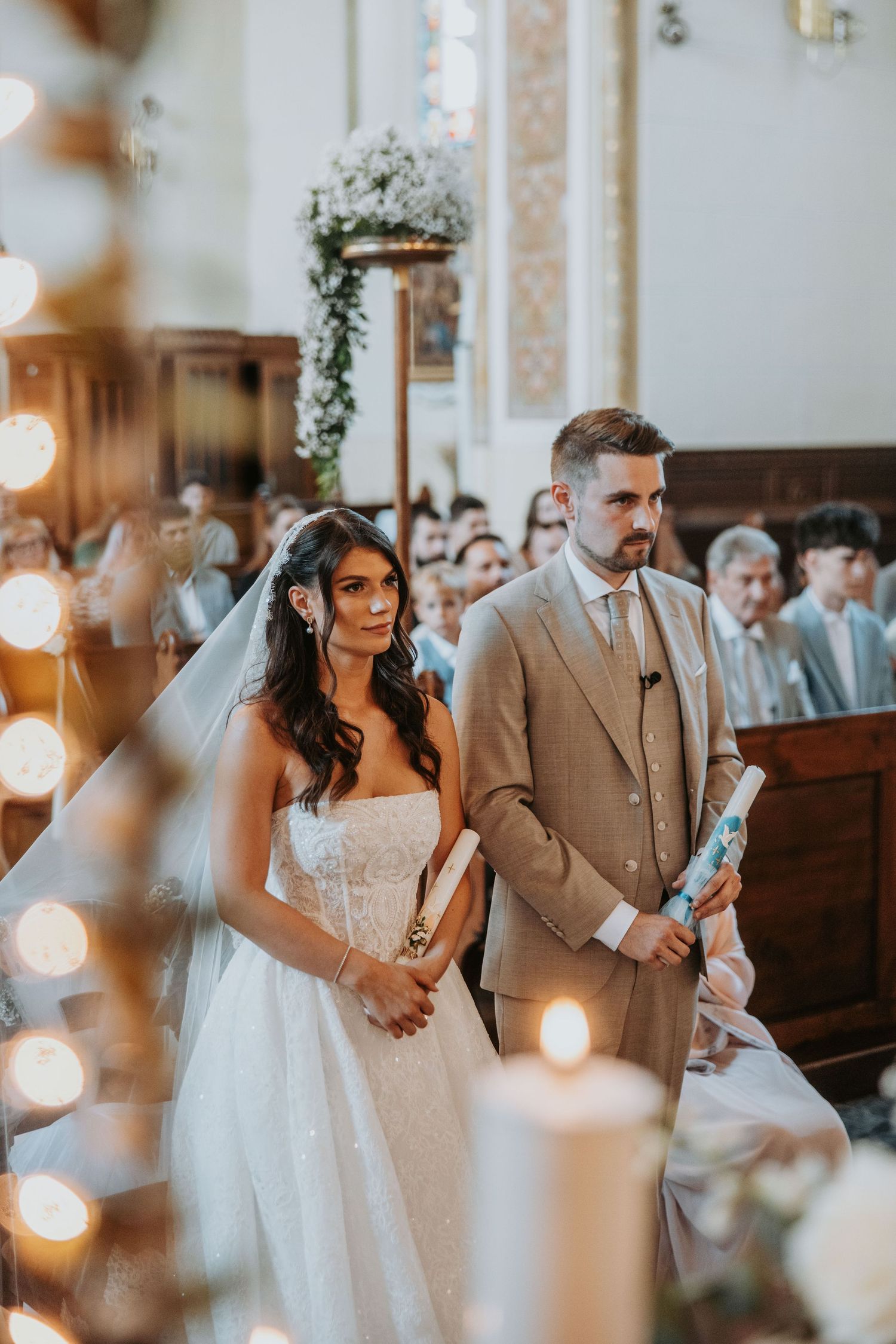 Bride and groom standing at church altar during wedding ceremony, bride in white gown with veil, groom in tan suit.