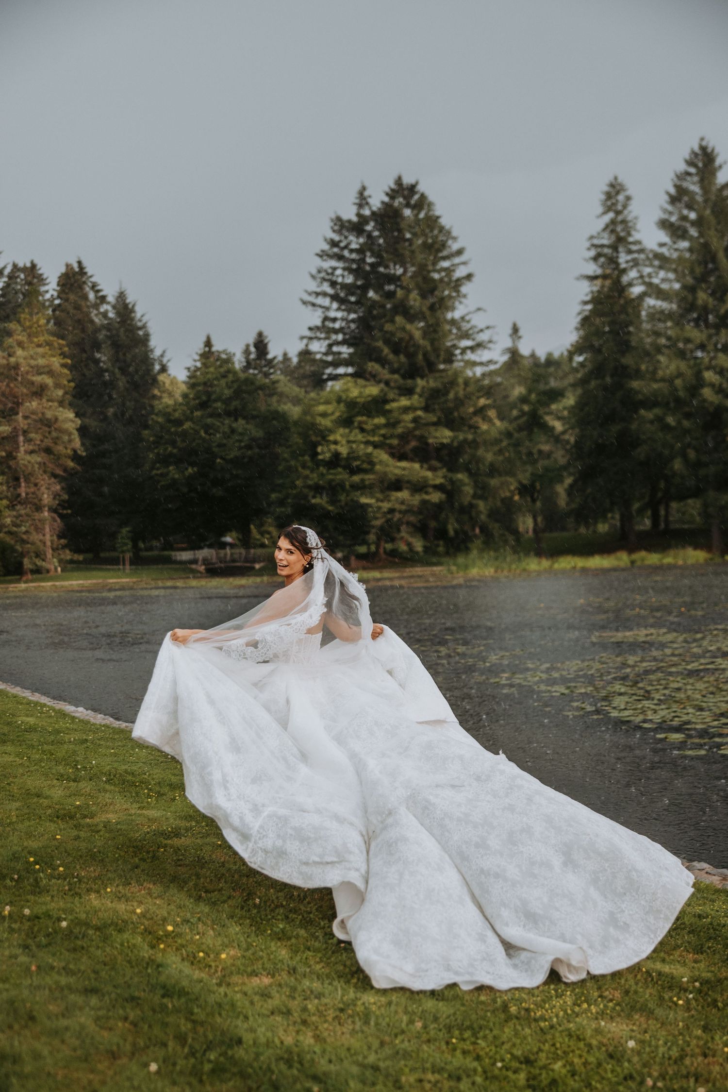 Bride in a flowing white lace gown with veil poses gracefully by a serene lake surrounded by tall evergreen trees.