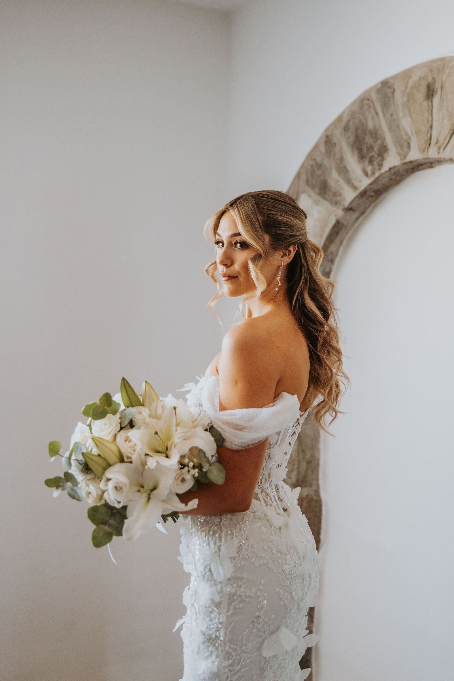 Bride in off-shoulder lace wedding gown holding white floral bouquet, posing elegantly beside stone arch.