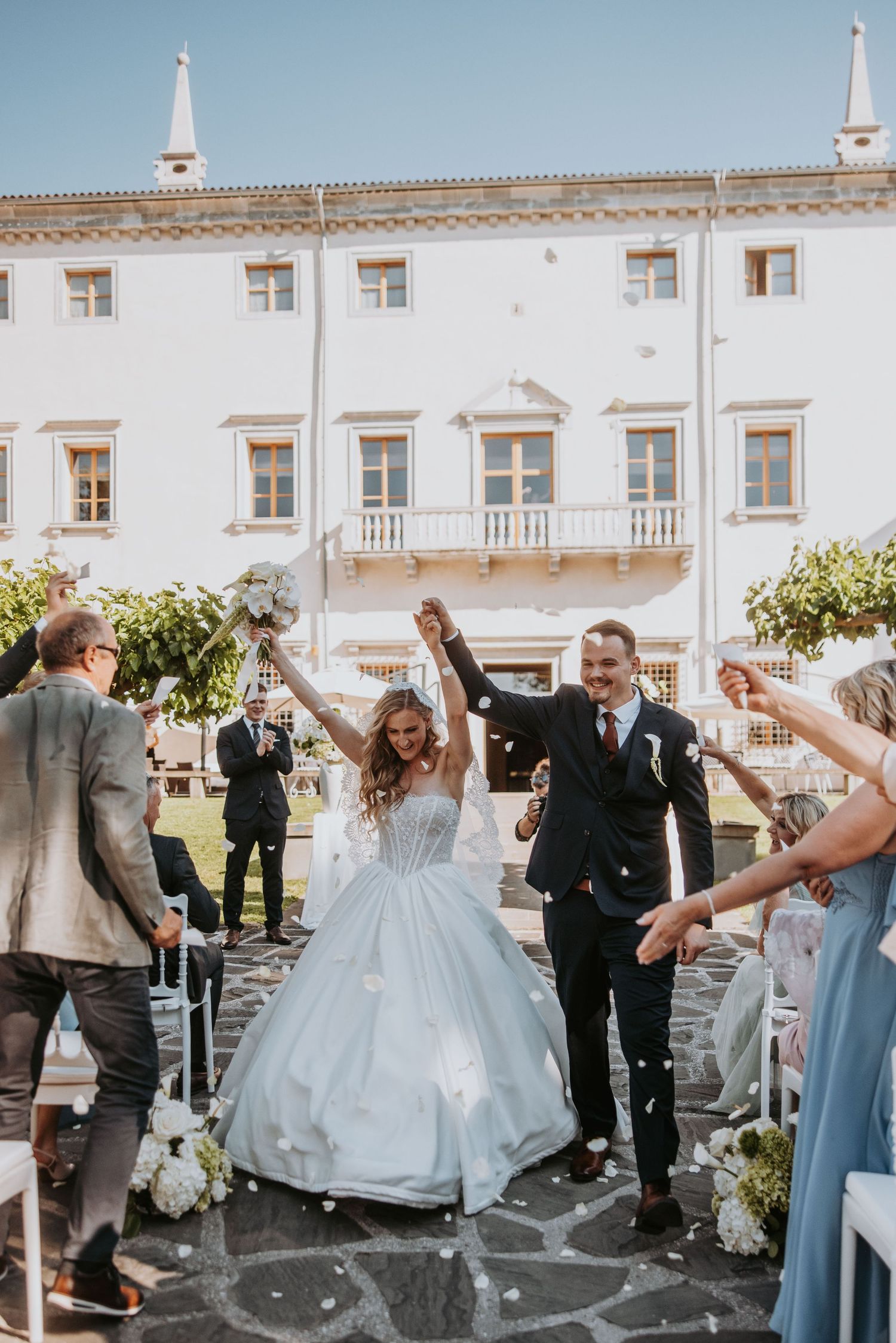 Joyful newlyweds celebrate with raised hands as guests toss confetti outside elegant white villa venue.
