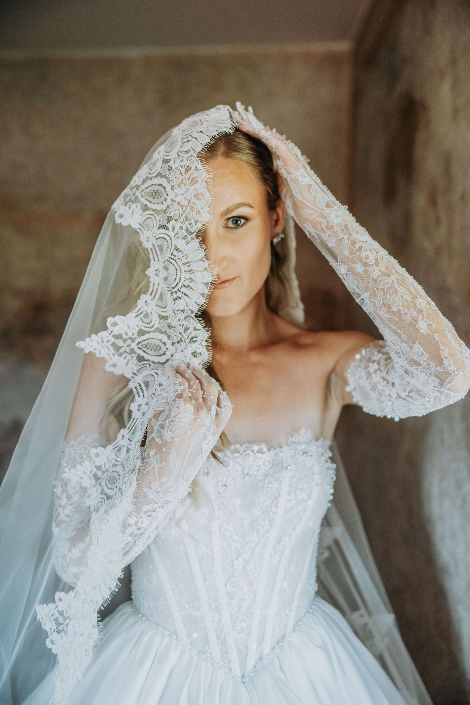 Elegant bride in lace wedding dress holds veil over face revealing beautiful embroidered details.