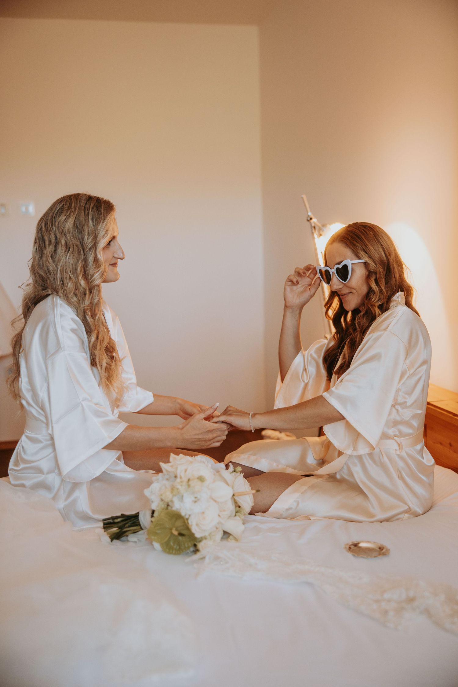 Two bridesmaids in white satin robes sit on a bed with white flowers during wedding preparations.
