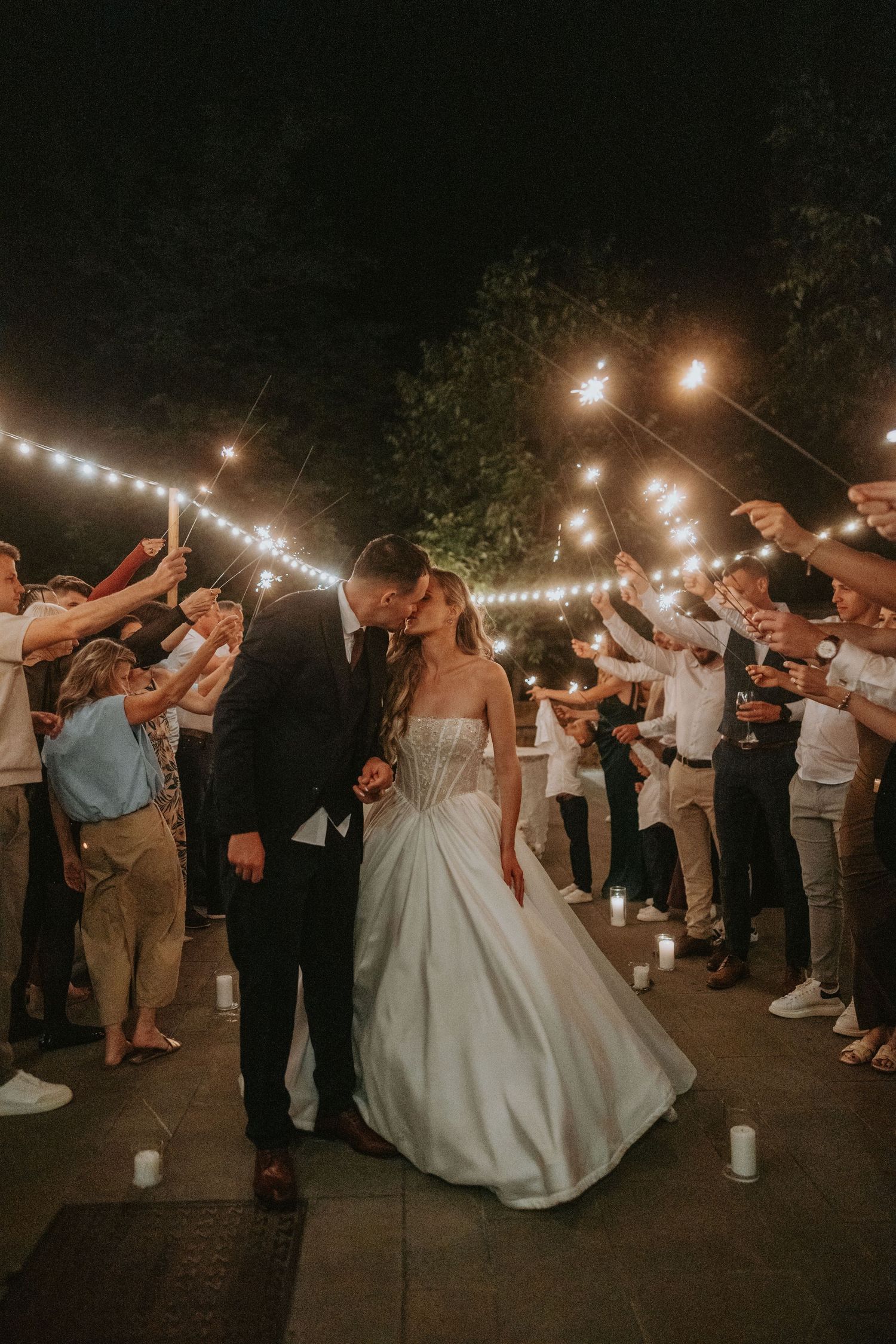 Bride and groom kiss during romantic sparkler send-off surrounded by guests at night wedding celebration.