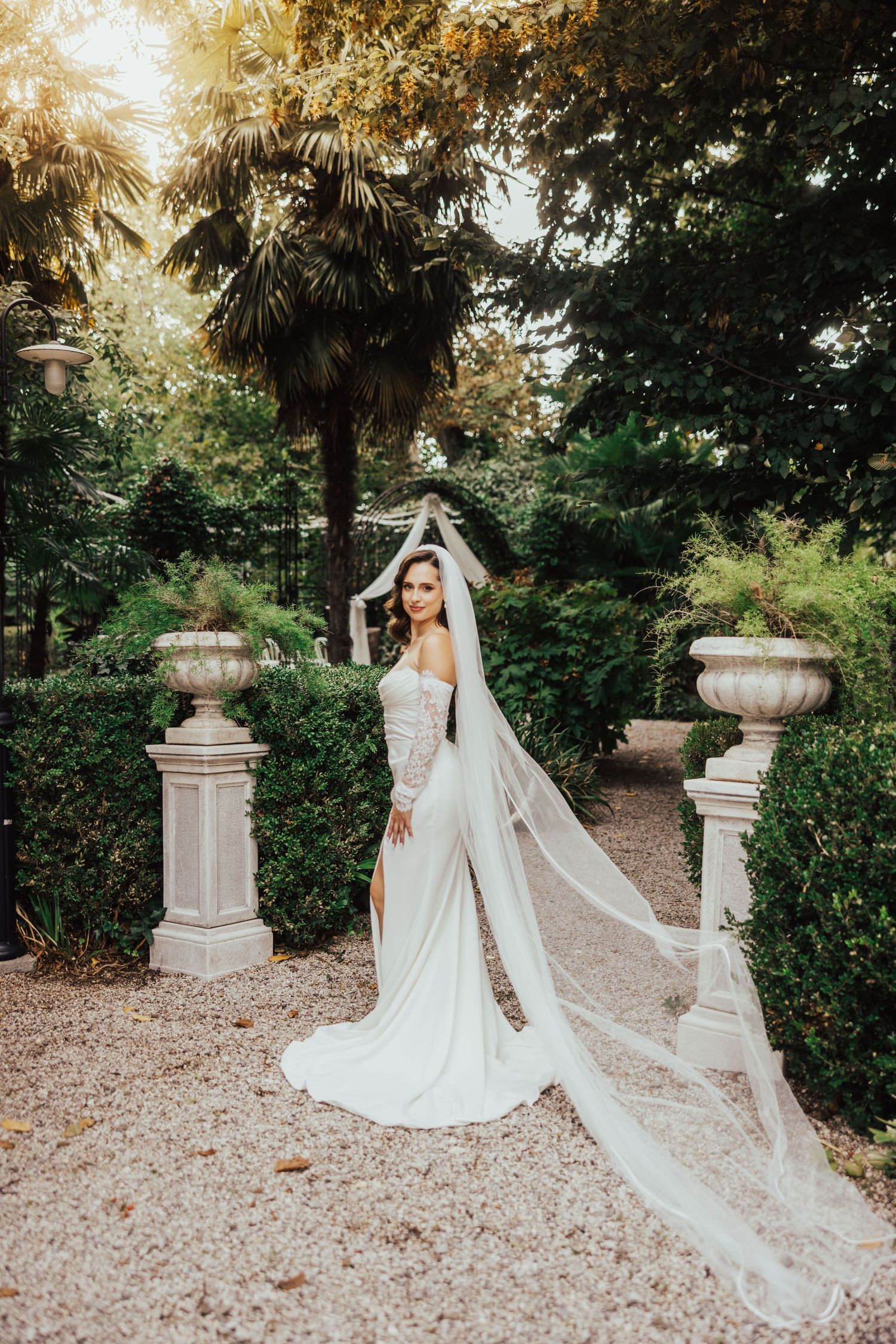 Bride in white off-shoulder gown with long flowing veil posing in lush garden with stone urns at sunset.