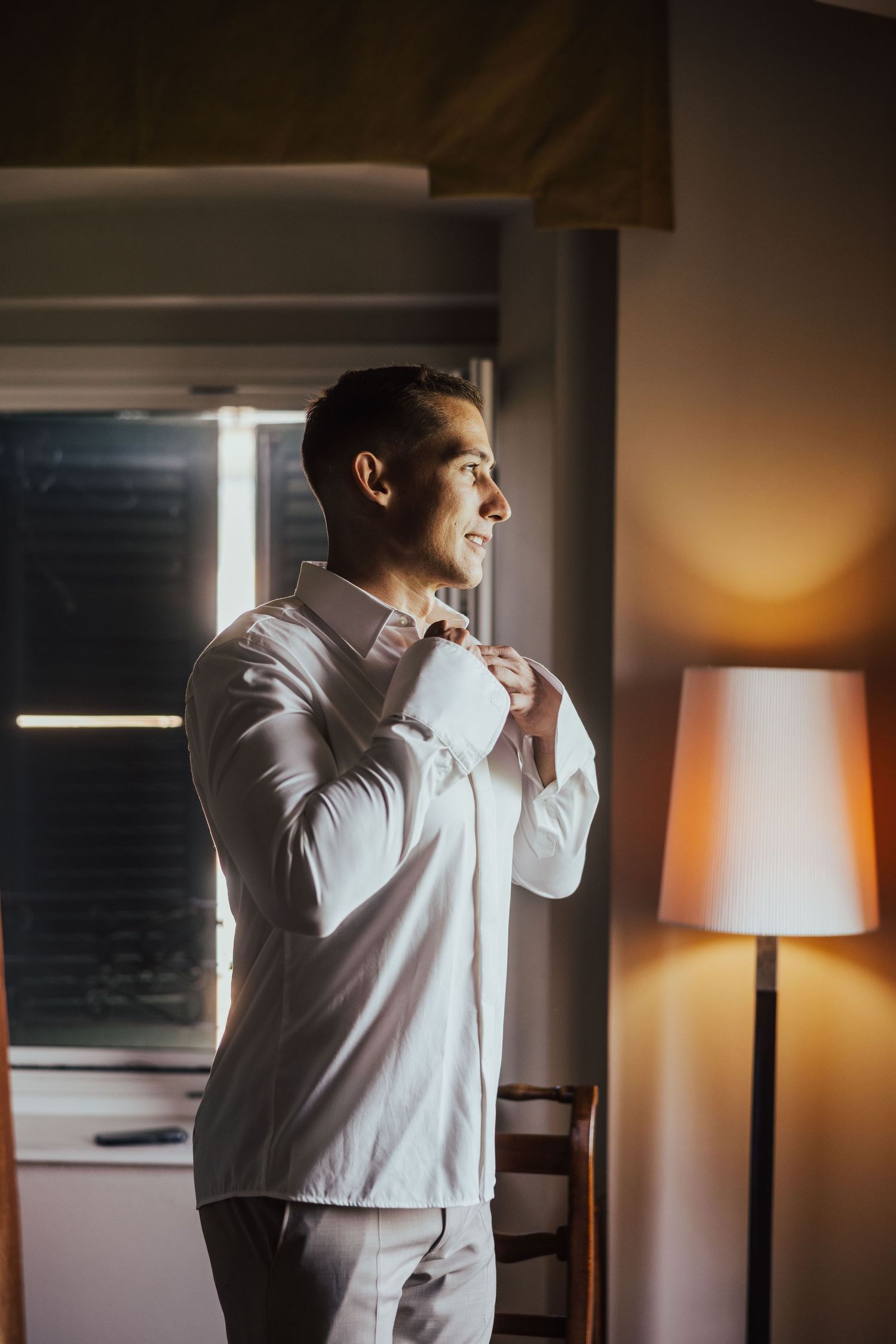 Groom adjusting his bow tie by a sunlit window in a white dress shirt before his wedding ceremony.