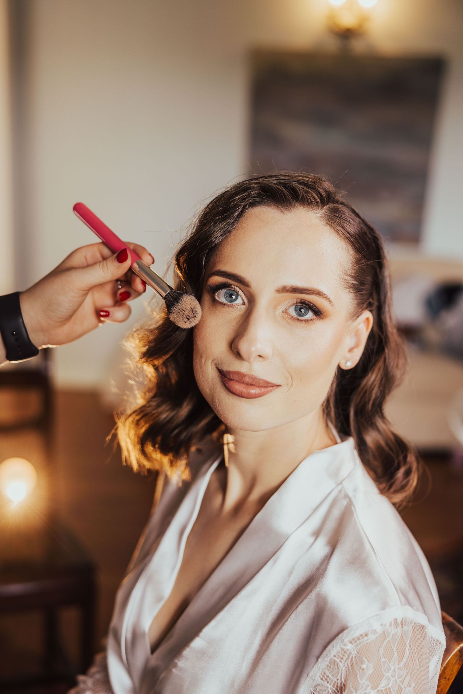 A makeup artist applies powder blush to a blue-eyed brunette bride in a white robe during wedding preparations.