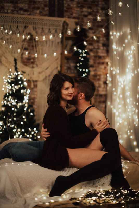 Couple embracing on a bed surrounded by Christmas tree lights and festive holiday decor.