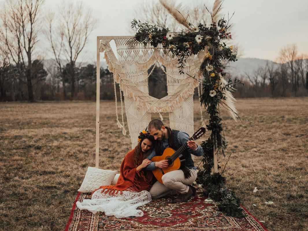 Couple sitting on a vintage rug under a boho macrame arch with flowers, man playing guitar in an open field.
