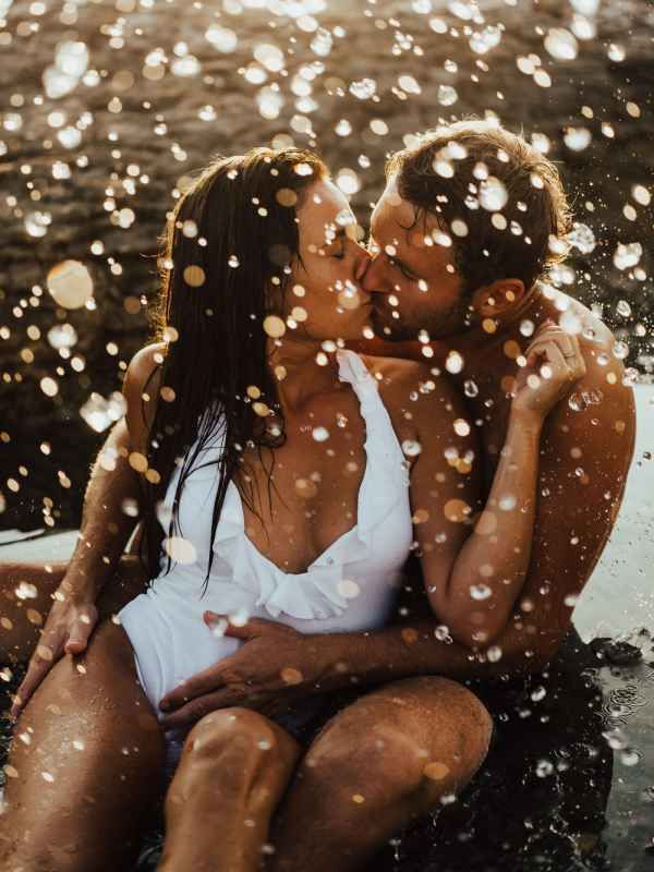 Couple kissing passionately on rocky shore with sparkling ocean water droplets surrounding them.