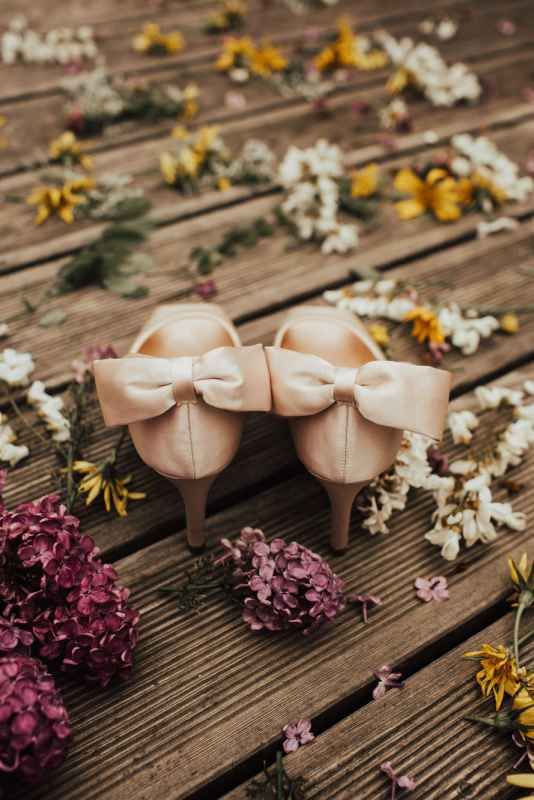 Elegant blush bridal heels with satin bows surrounded by colorful wildflowers on wood.