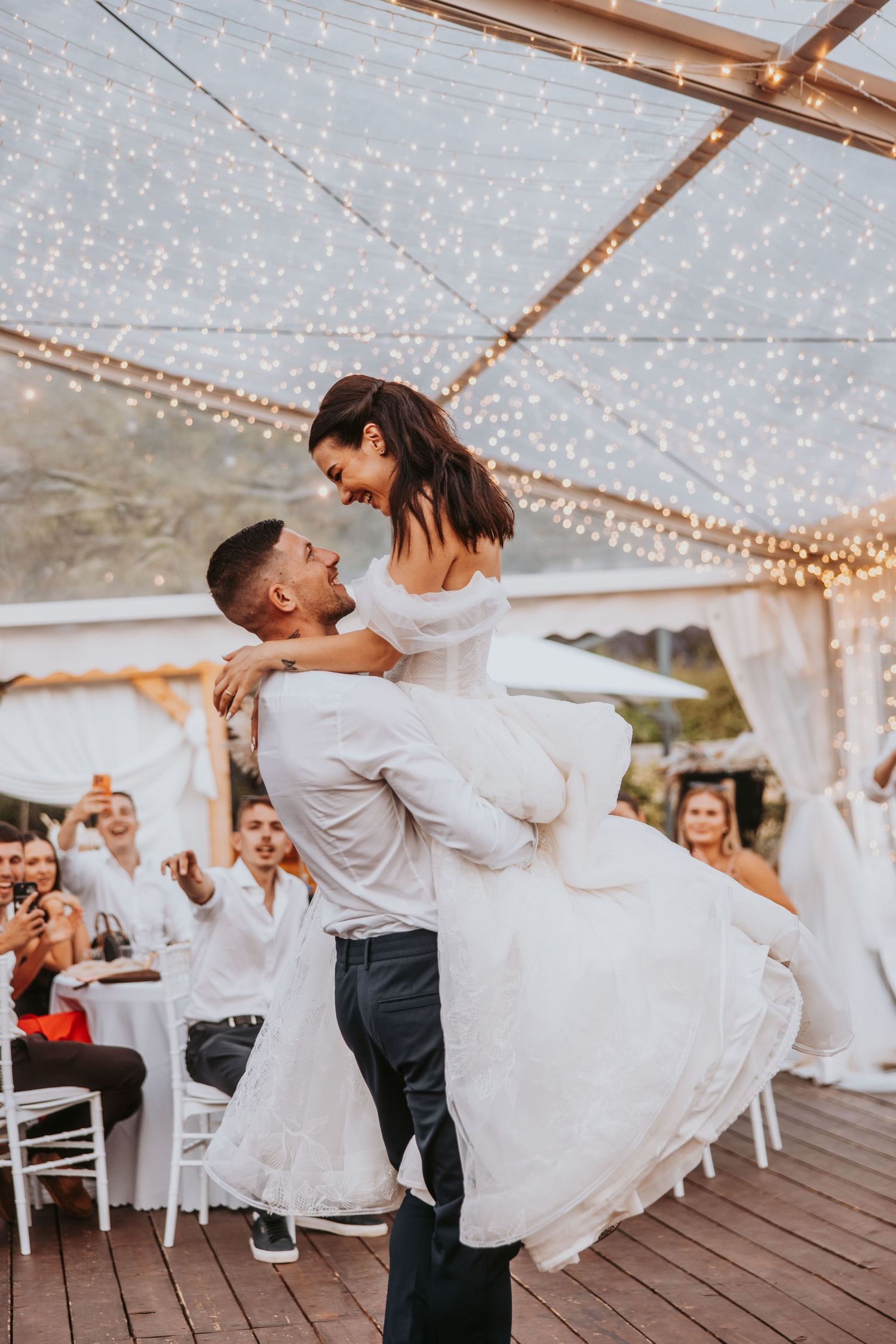 Groom lifting bride in white dress during first dance under fairy lights at wedding reception.