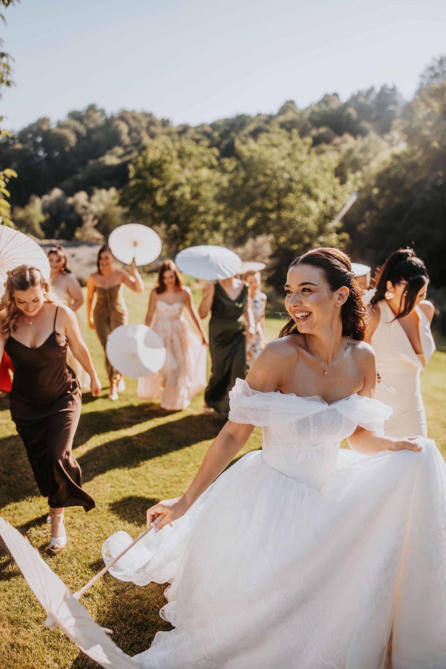 Joyful bride in white off-shoulder gown running outdoors with guests holding parasols on sunny wedding day.