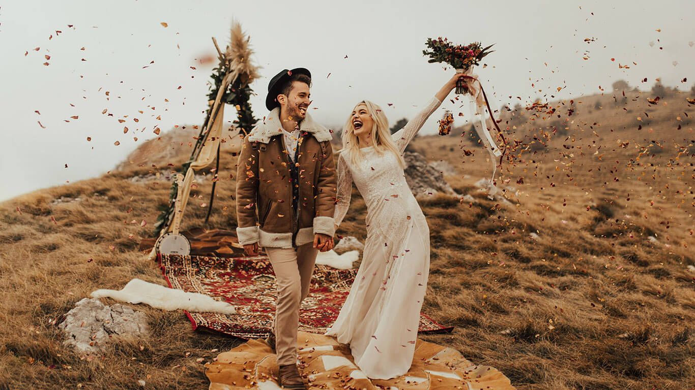 Boho bride and groom celebrating on a hillside with flower petals and rustic decorations.