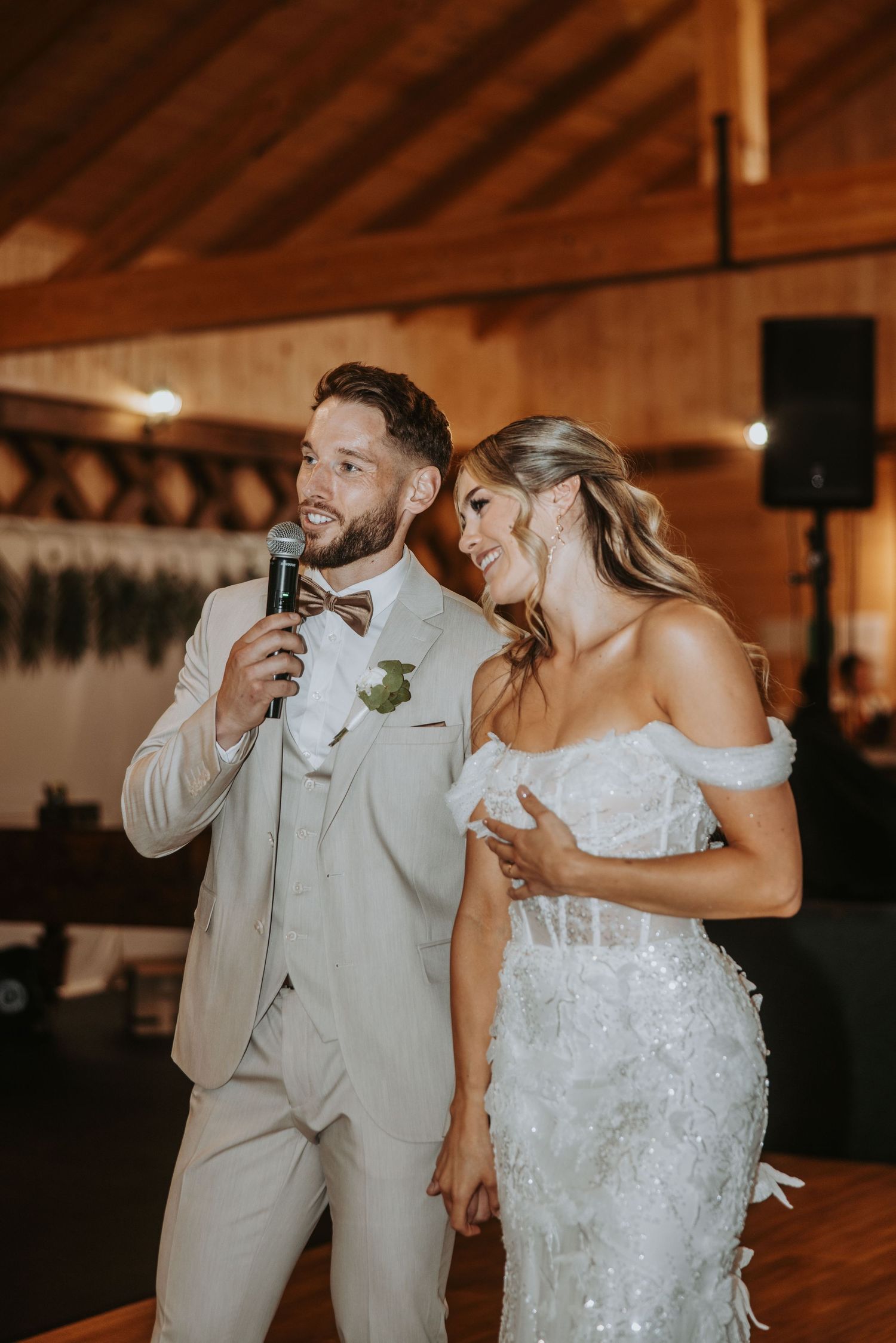 Groom speaking into microphone at wedding reception while bride laughs beside him in rustic venue.
