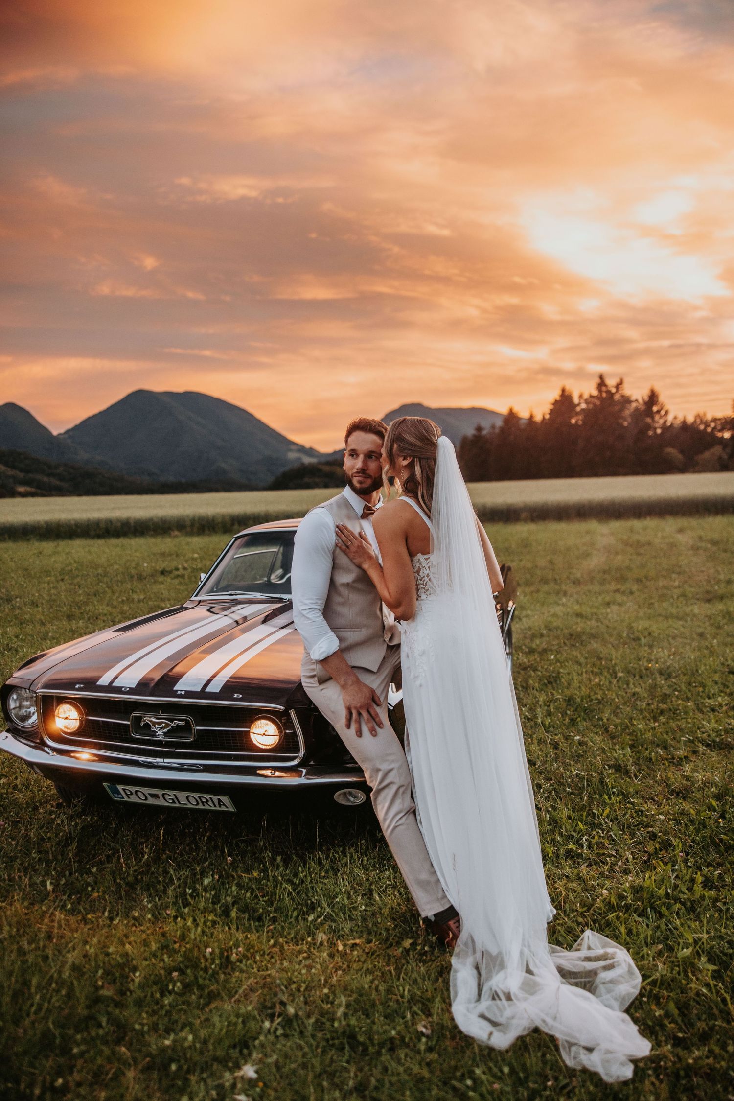 Bride and groom pose against vintage Ford Mustang in field during stunning golden sunset.
