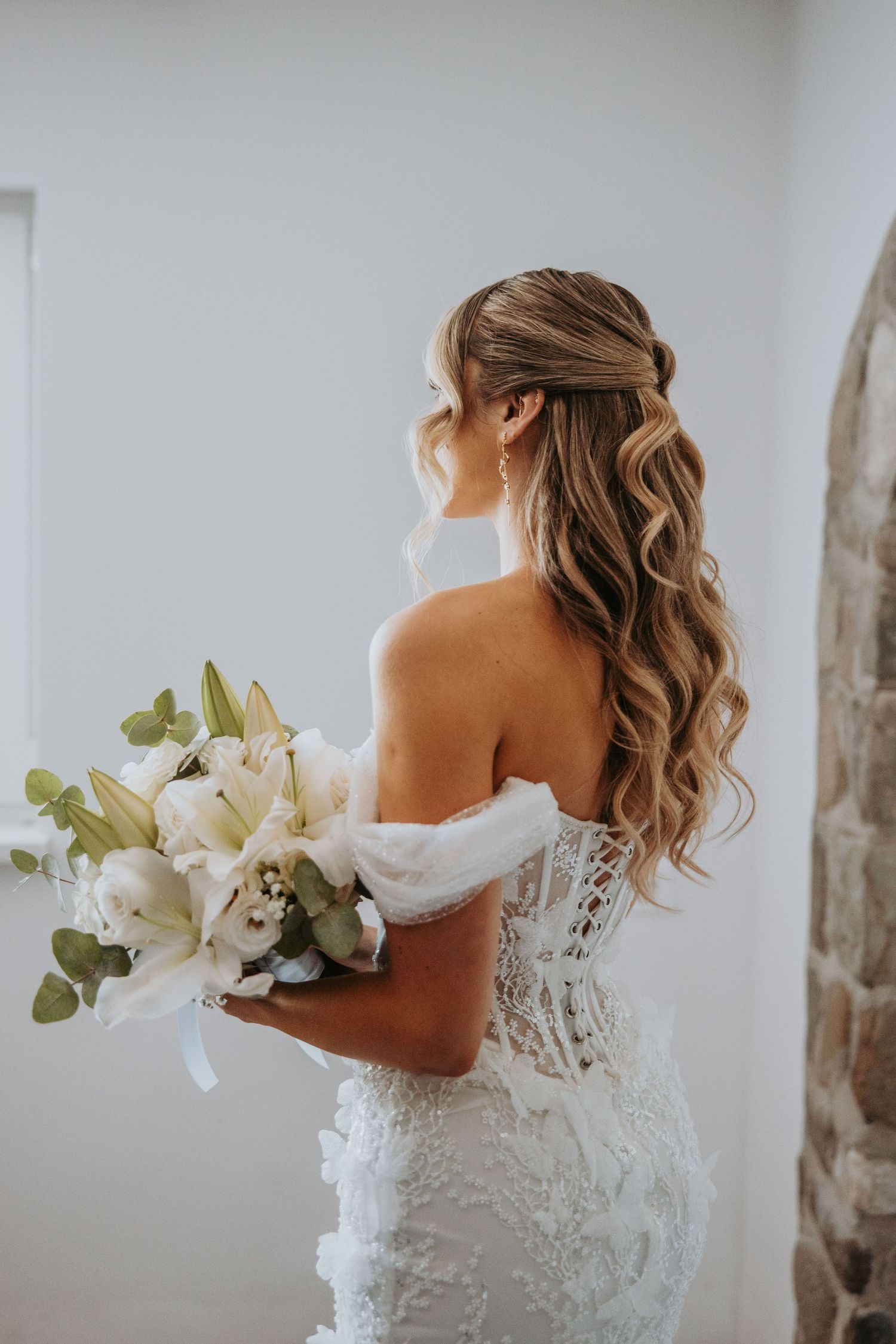 Elegant bride in lace off-shoulder wedding gown holding white lily bouquet, showcasing curled half-up hairstyle.