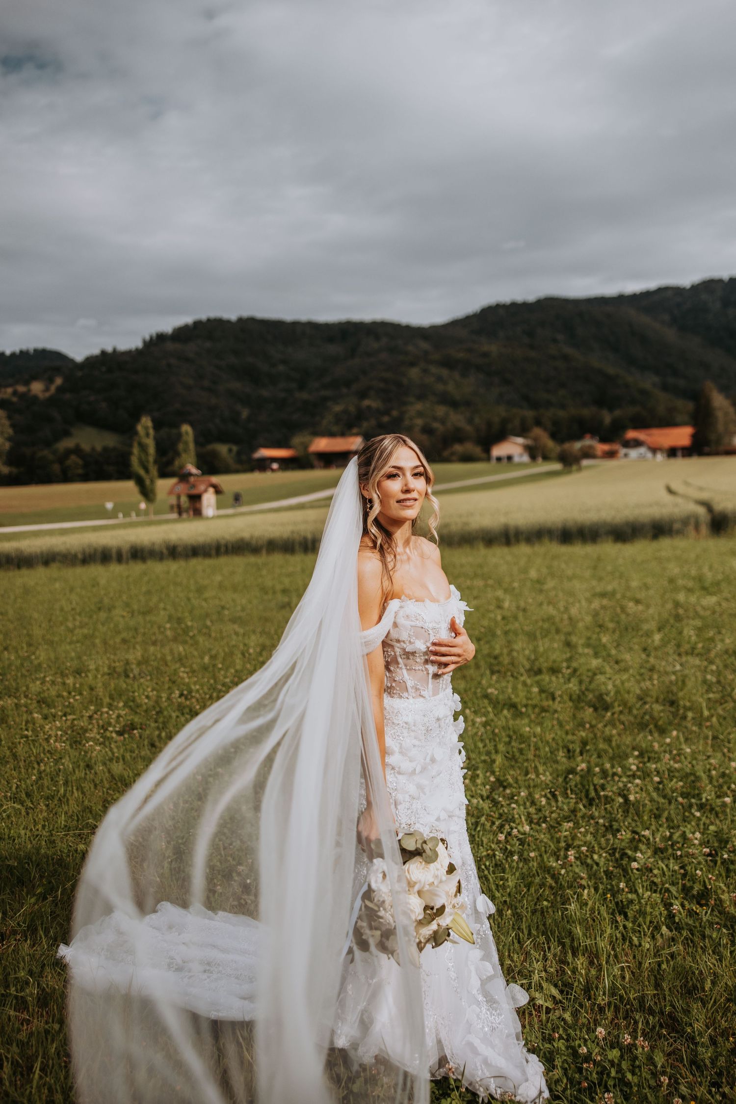 Bride in floral lace wedding dress with veil blowing in the wind standing in a green mountain meadow.