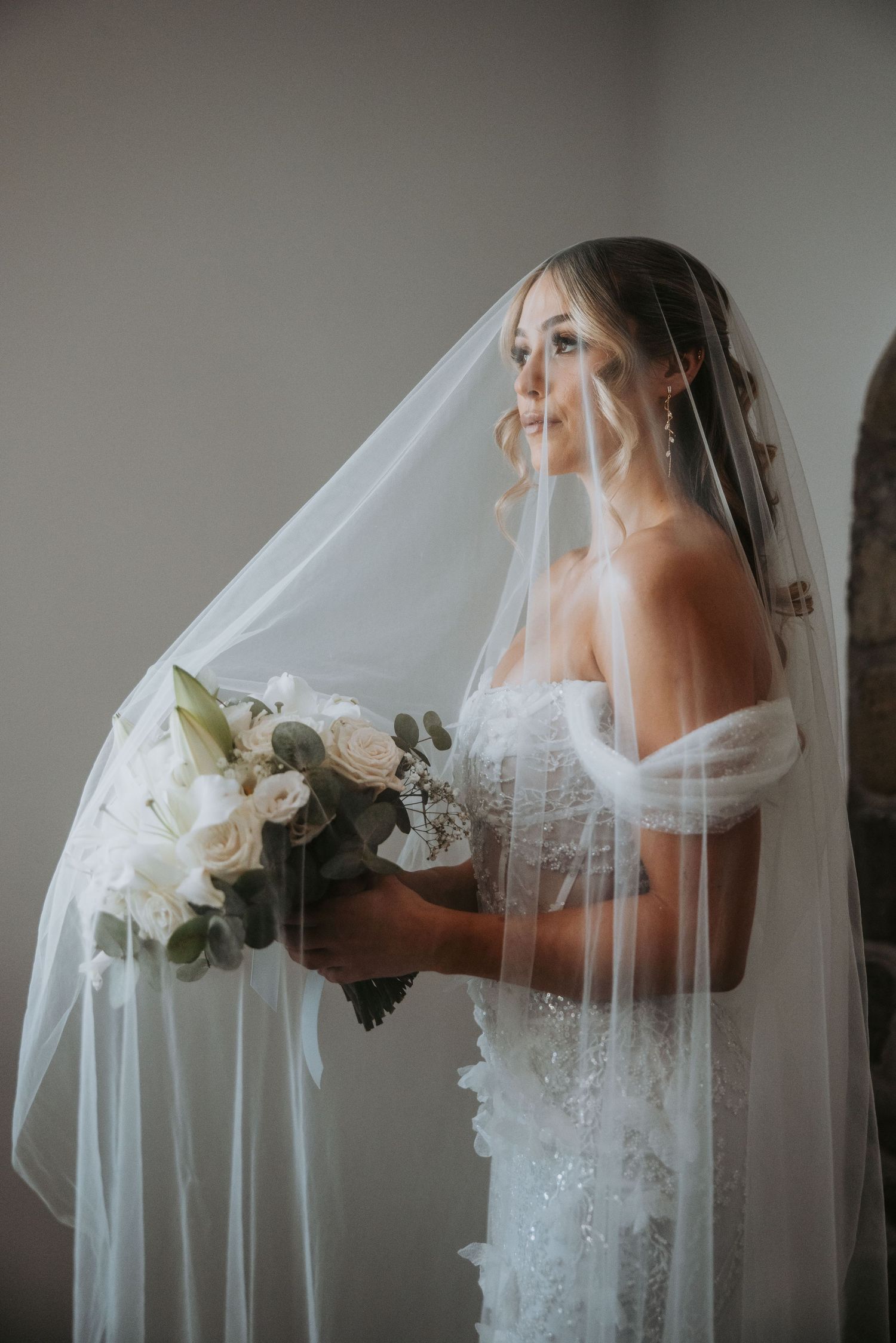 Bride in lace off-shoulder gown holds white floral bouquet under flowing veil in soft natural light.