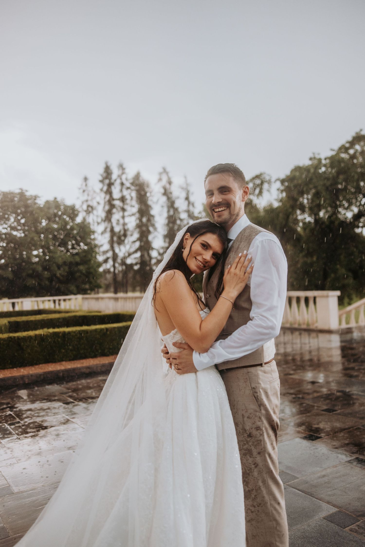 Smiling bride and groom embrace tenderly in the rain during their outdoor wedding portrait session.