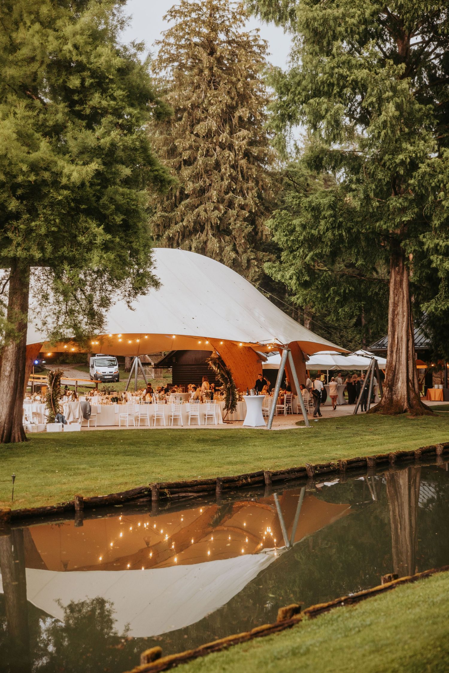 Elegant outdoor wedding reception tent with warm string lights reflected in a pond surrounded by tall trees at dusk.