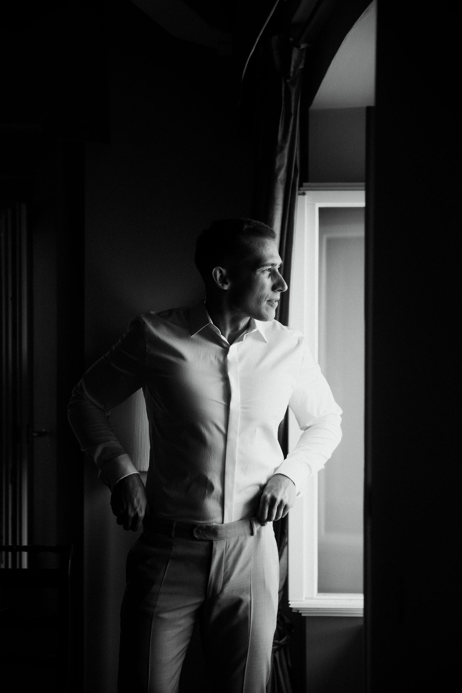 Black and white portrait of groom in white dress shirt standing by window before wedding ceremony.