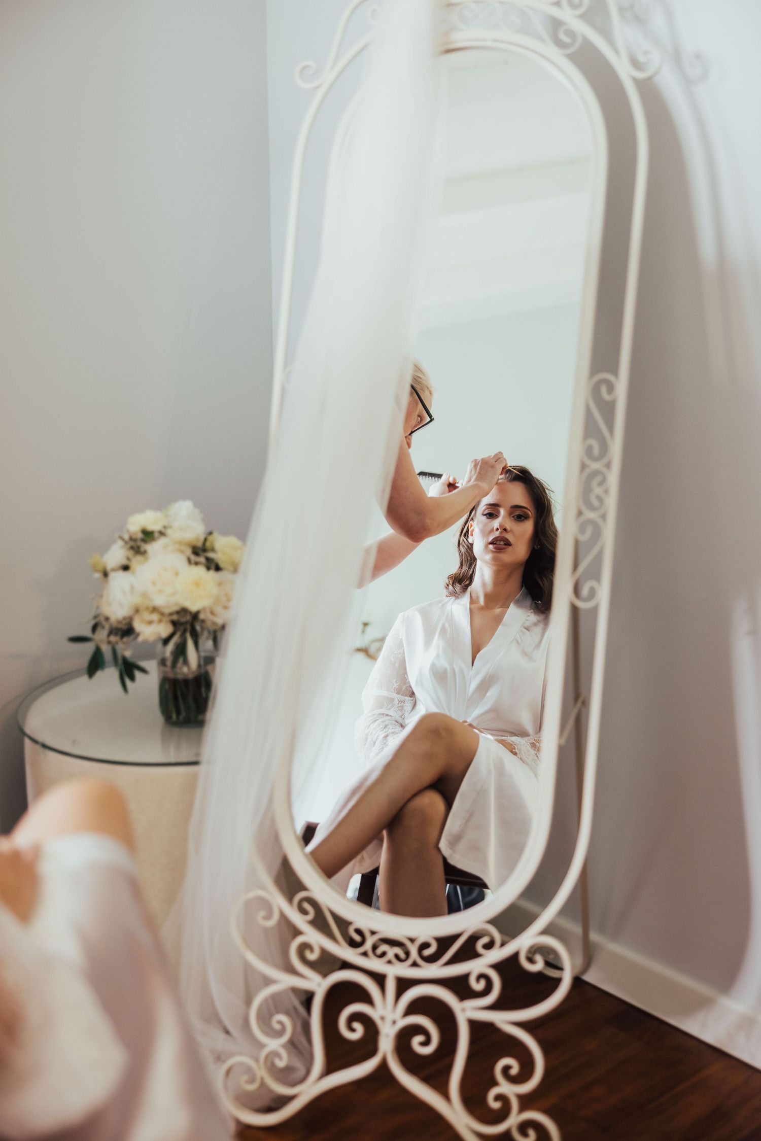 Bride in a white robe gets makeup applied while seated before an ornate white mirror in a bright bridal suite.