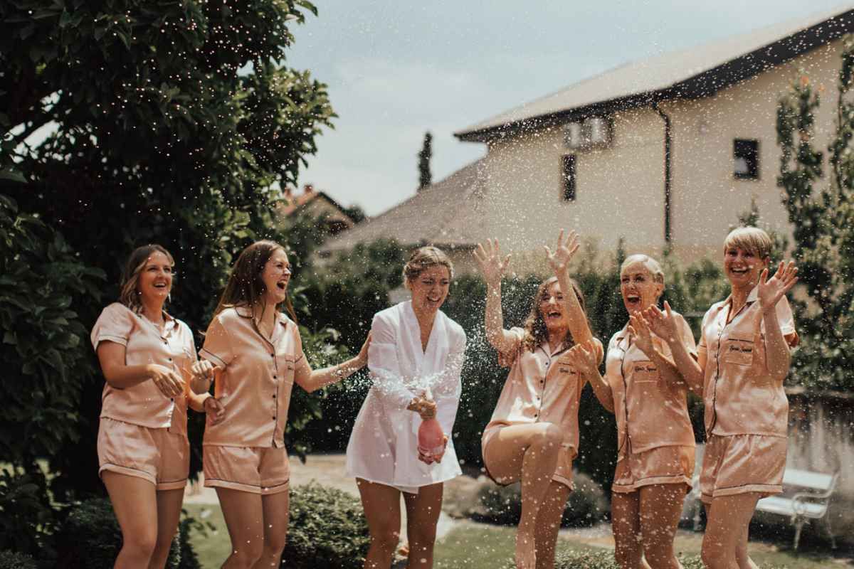 Bridesmaids in matching satin pajamas celebrate with champagne spray outdoors at a wedding morning party.