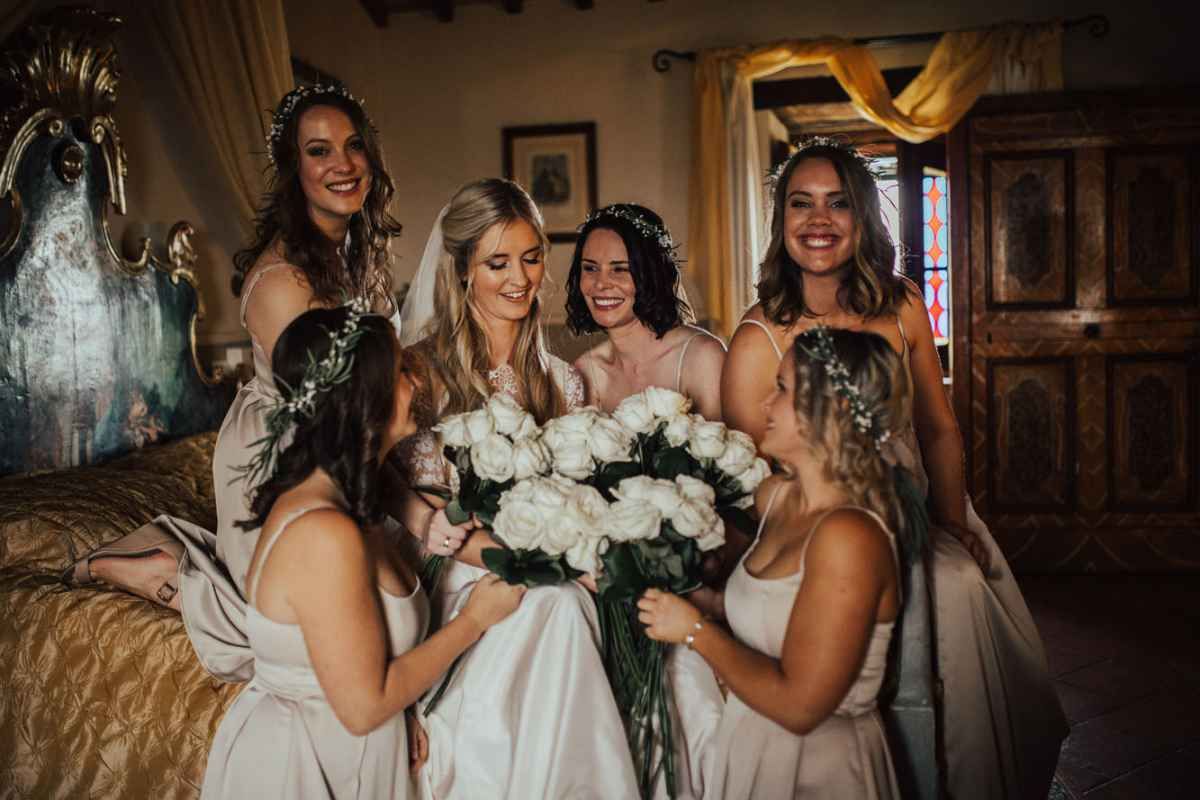 Bride holding white roses bouquet surrounded by smiling bridesmaids in champagne dresses inside elegant venue.