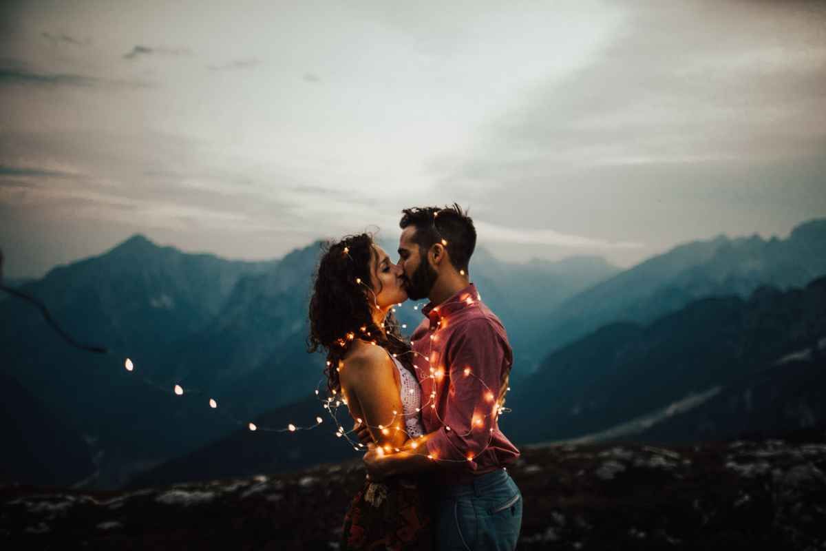 Romantic couple kissing wrapped in fairy lights with mountain backdrop at dusk.