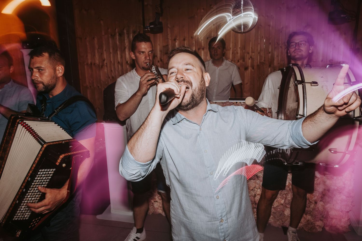 Energetic male singer performs with accordion band under colorful lights at lively wedding party.