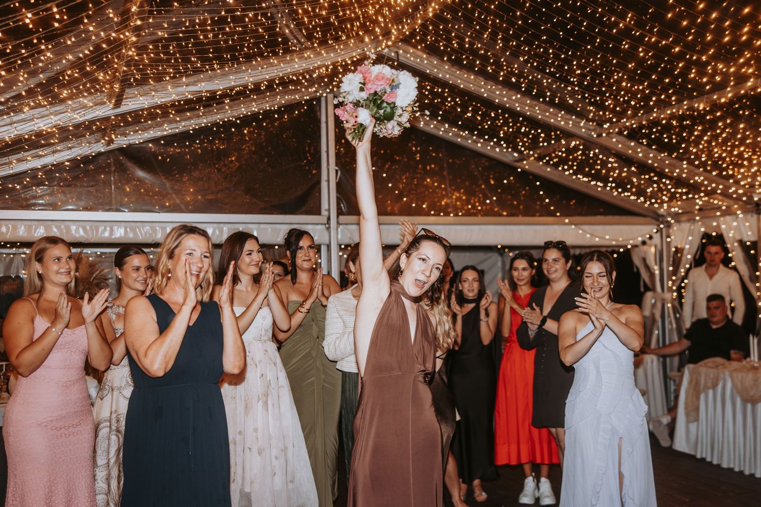 Excited wedding guest catches bridal bouquet toss under sparkling fairy lights at reception.