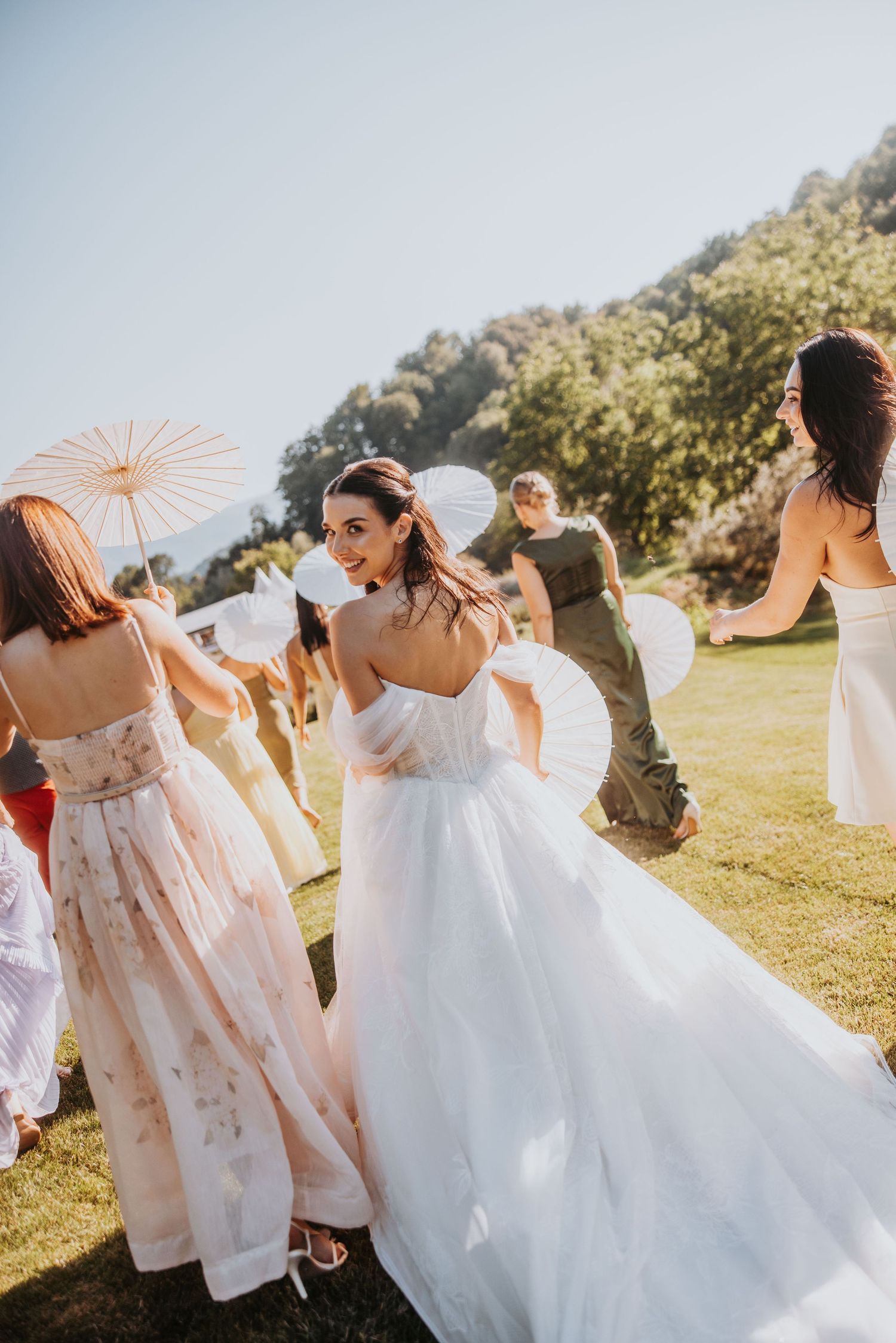 Bride in strapless white ballgown looking back with bridesmaids carrying paper parasols at sunny outdoor wedding.
