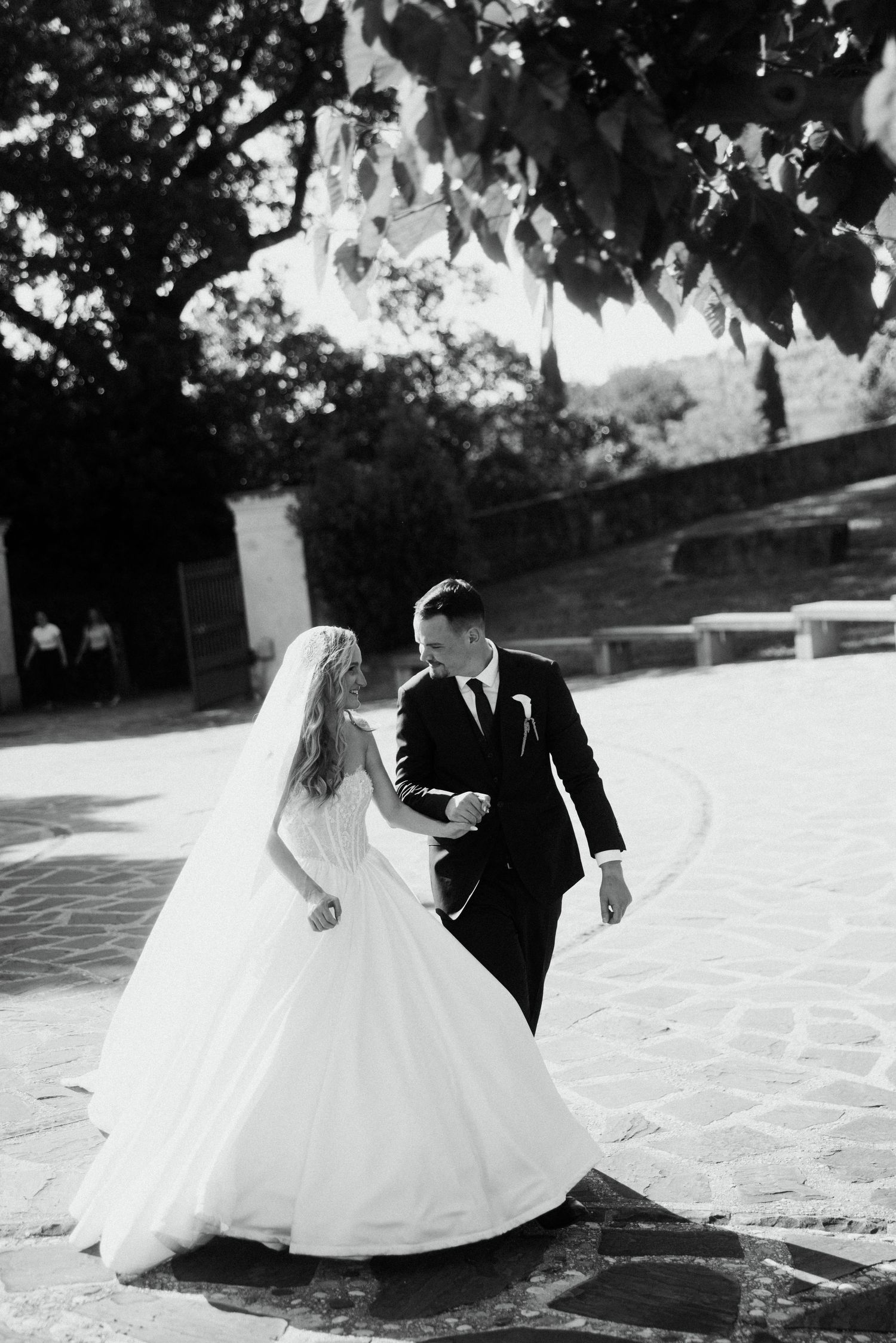 Black and white photo of bride and groom walking hand in hand on stone courtyard under tree canopy.