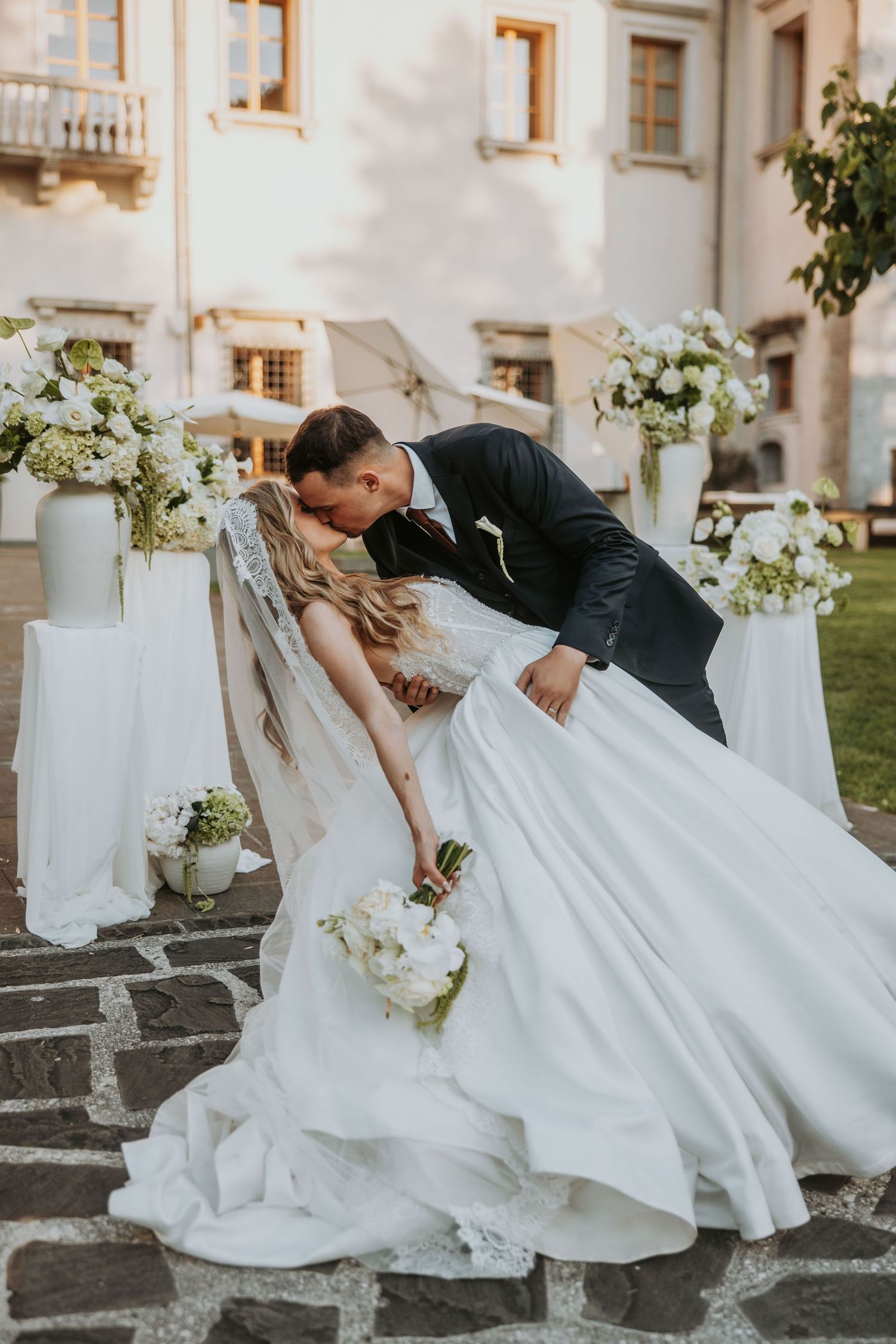 Groom dipping bride for romantic kiss at outdoor wedding ceremony with white floral arrangements.