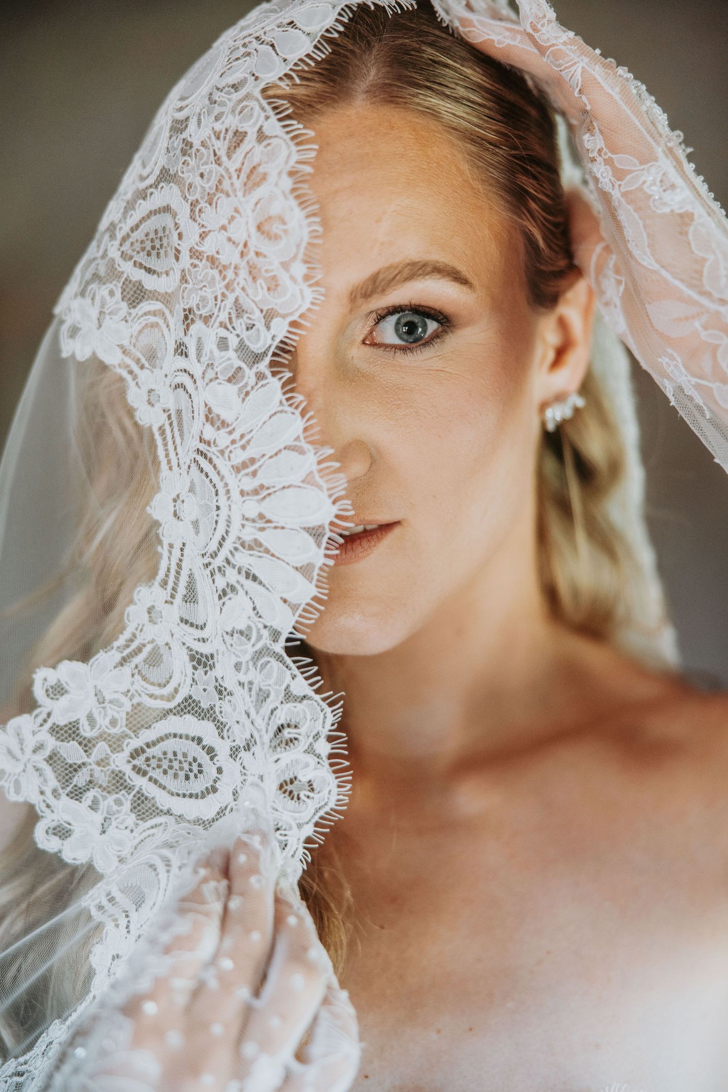 Bride holding lace veil over half her face, revealing blue eyes and pearl earring in elegant bridal portrait.