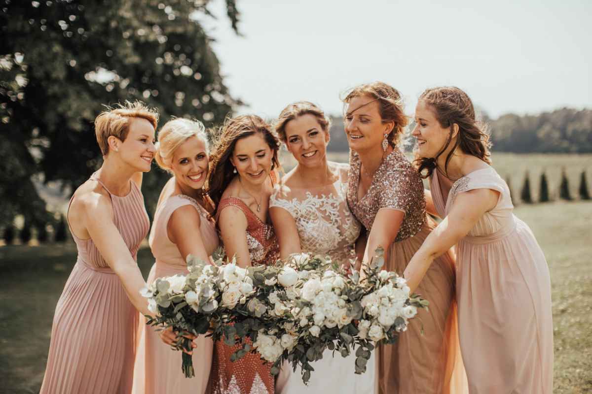 Bride and bridesmaids in pink dresses laughing together holding white floral bouquets outdoors.