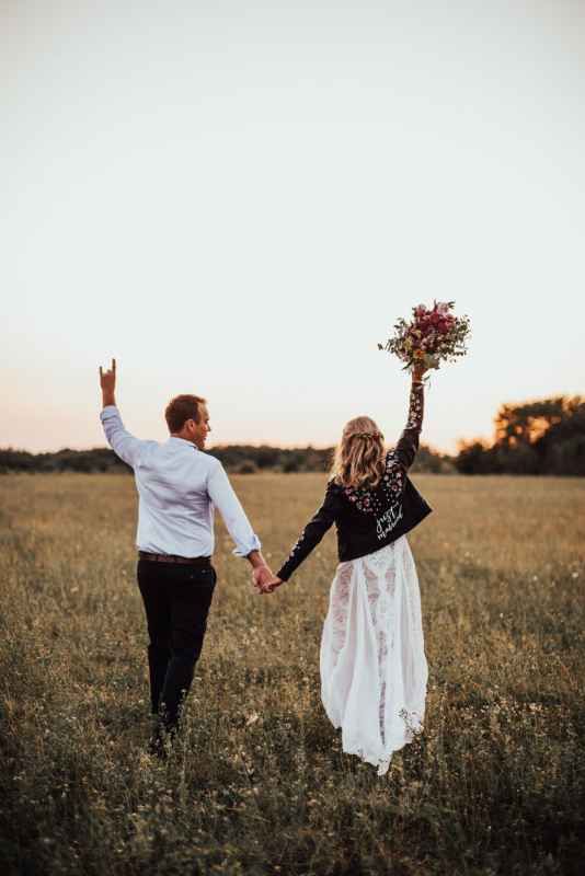 Wedding couple holding hands in golden field at sunset, bride in lace dress and leather jacket.