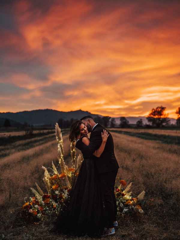 Couple embracing in a field at sunset with vibrant orange sky and floral arrangement at golden hour.
