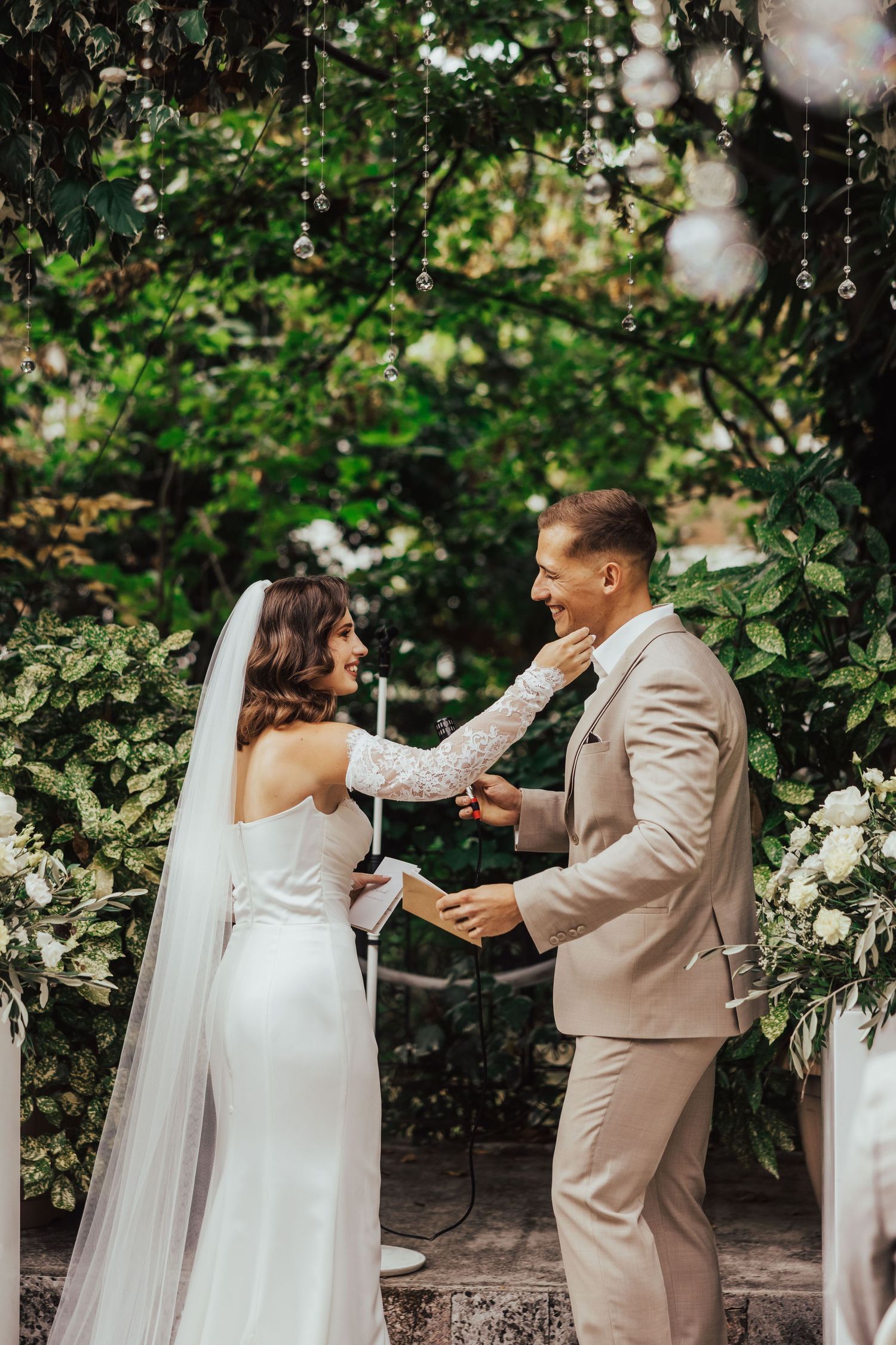 Bride and groom exchange vows at outdoor wedding ceremony surrounded by lush greenery and white floral arrangements.