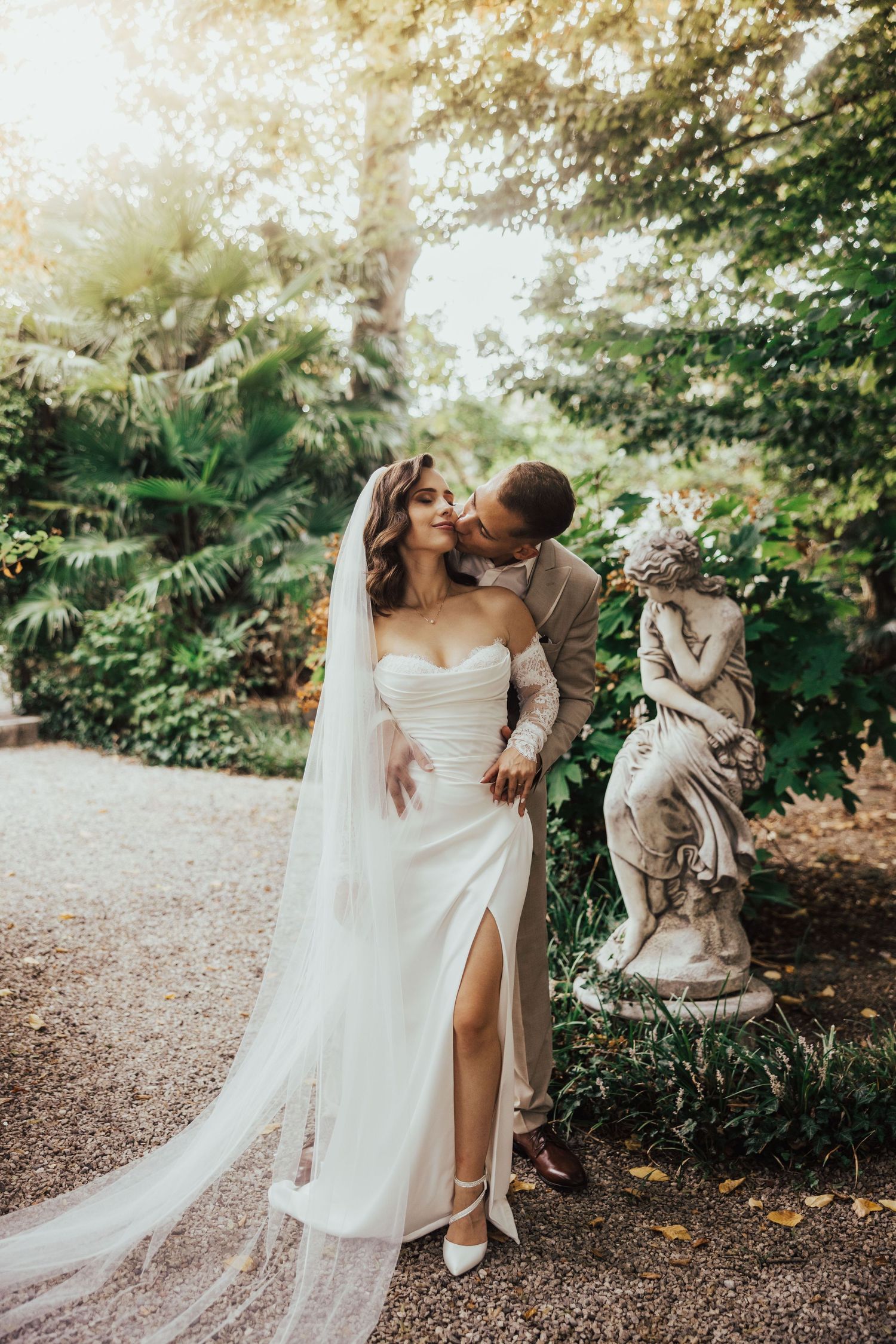 Elegant bride in a strapless white gown with a long veil stands with groom in a garden beside a stone cherub statue.