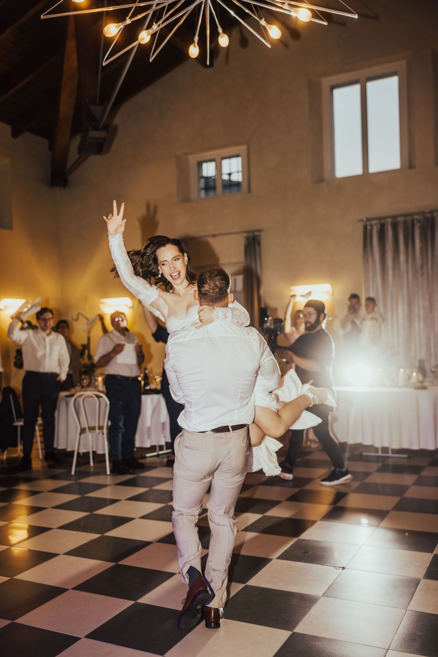 Groom lifts joyful bride during energetic first dance on checkered floor at wedding reception.