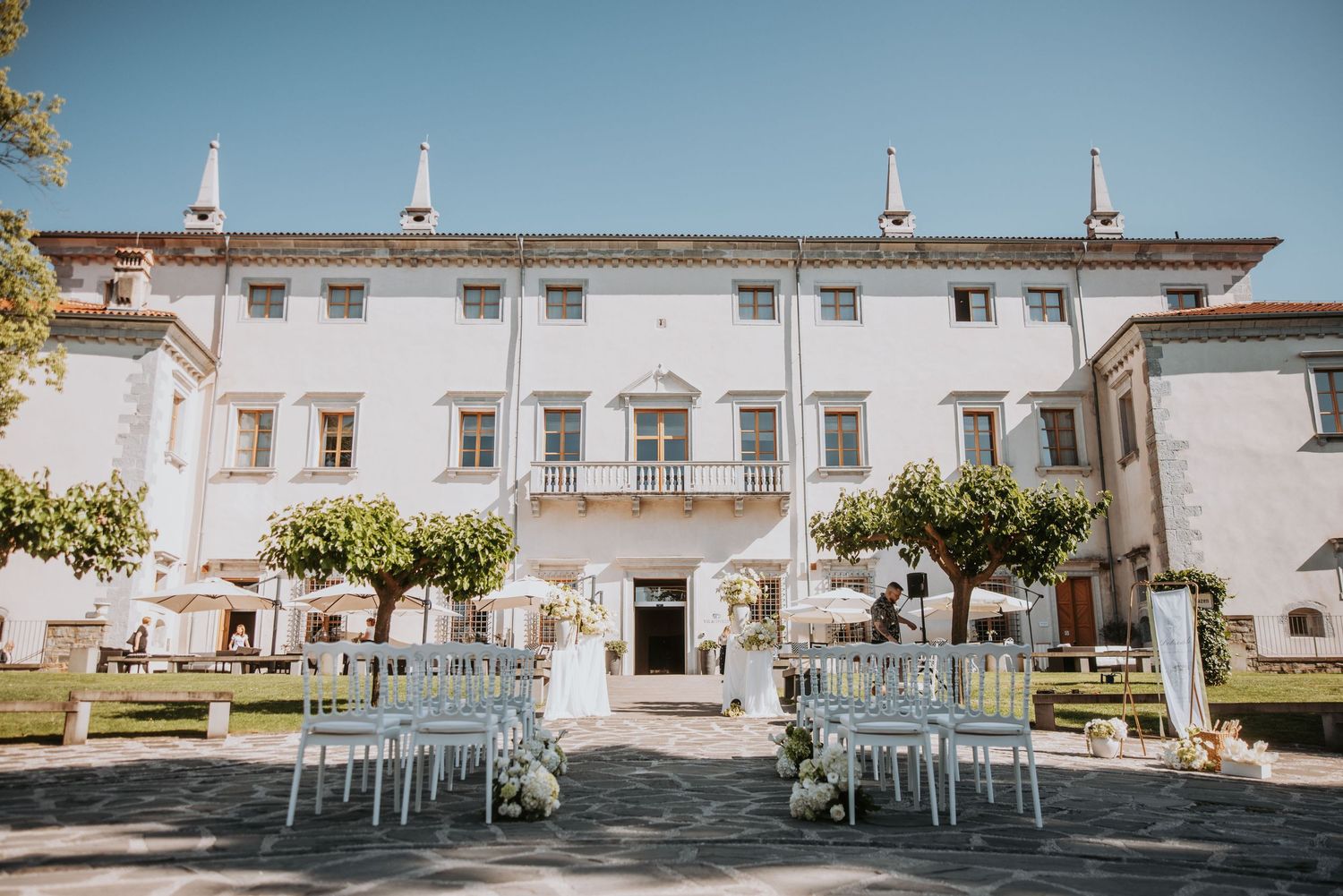 Elegant white villa wedding venue with outdoor seating arrangement, lush green trees, and blue chairs under clear sky.
