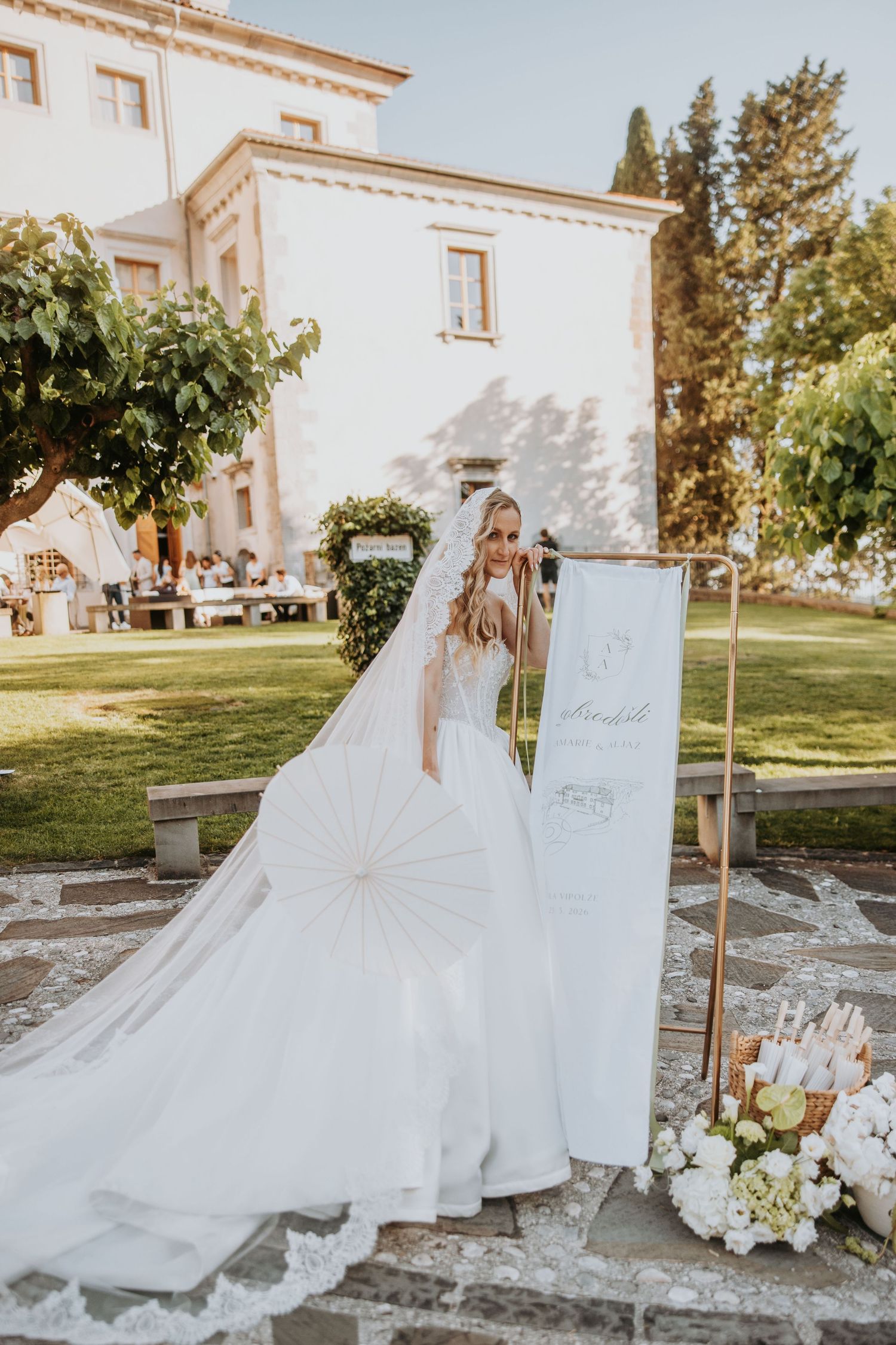 Elegant bride in a ball gown holds a parasol beside a welcome sign at a Tuscan villa wedding venue.